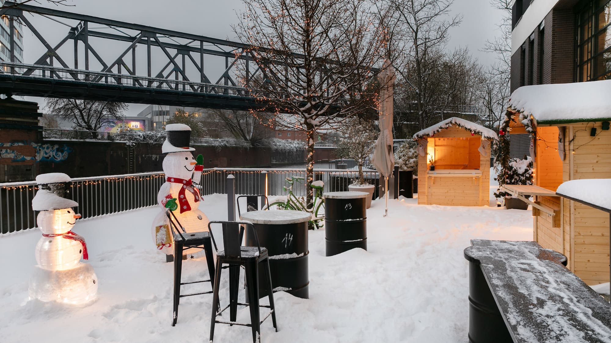 Snowy Christmas market with illuminated snowmen, festive stalls, and bridge.