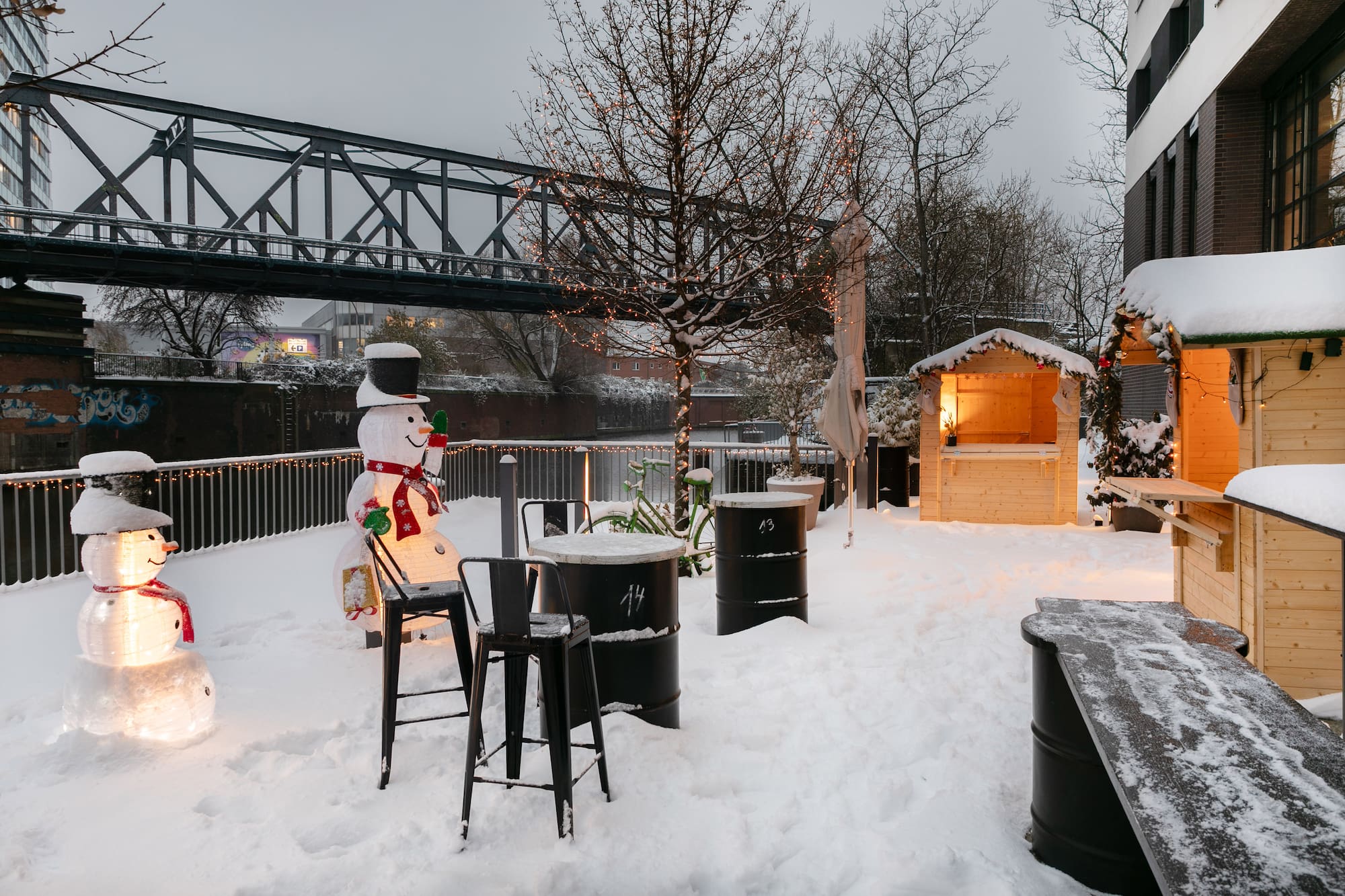 Snowy Christmas market with illuminated snowmen, festive stalls, and bridge.