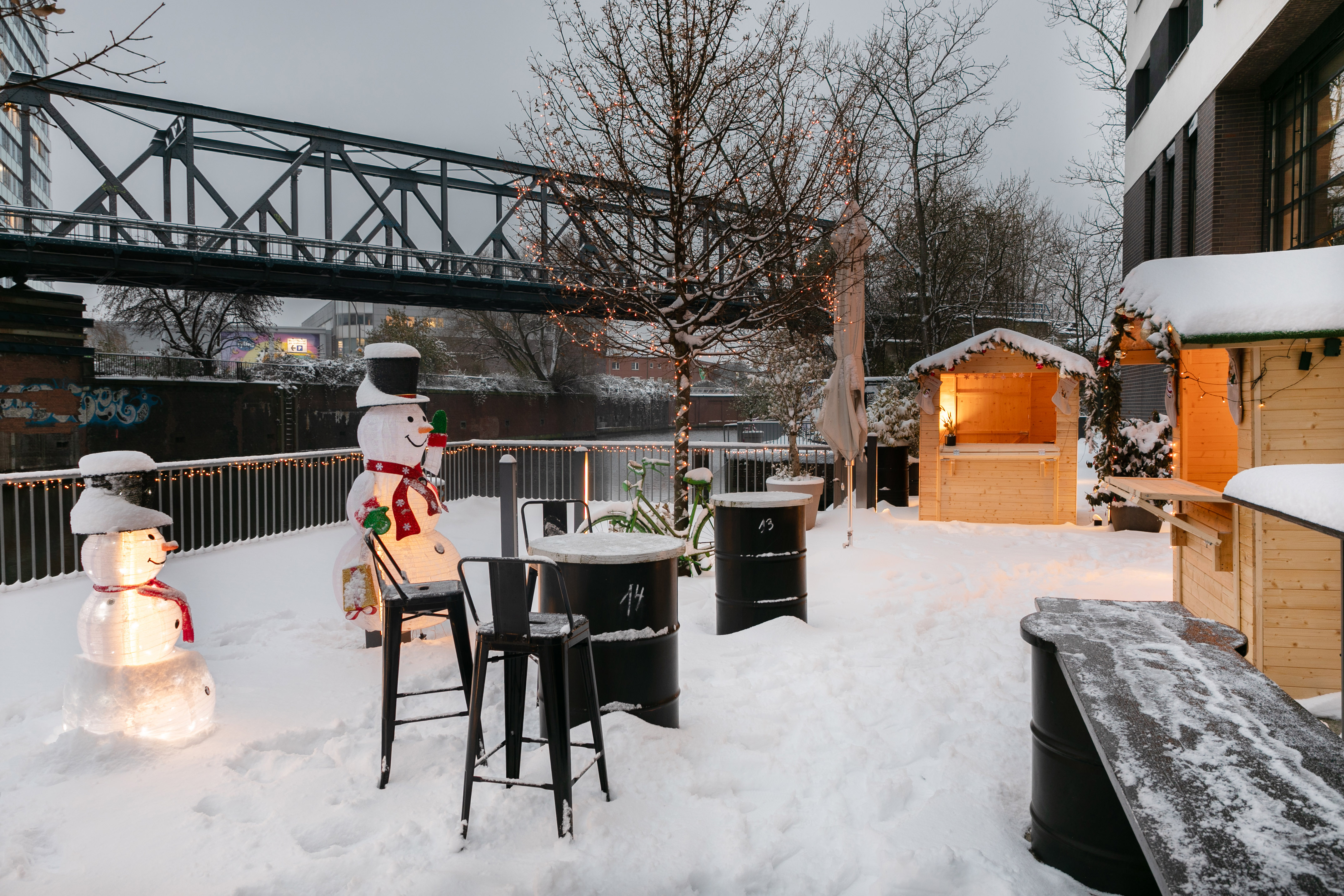 Snowy Christmas market with illuminated snowmen, festive stalls, and bridge.