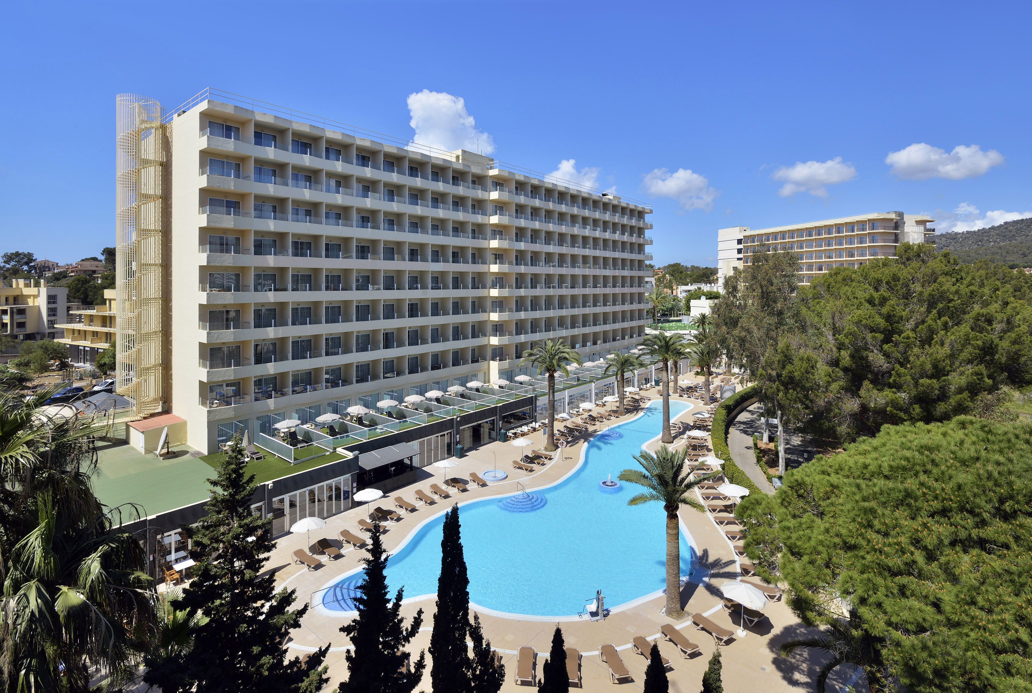 Sunny resort hotel with large blue pool, palm trees, and loungers.