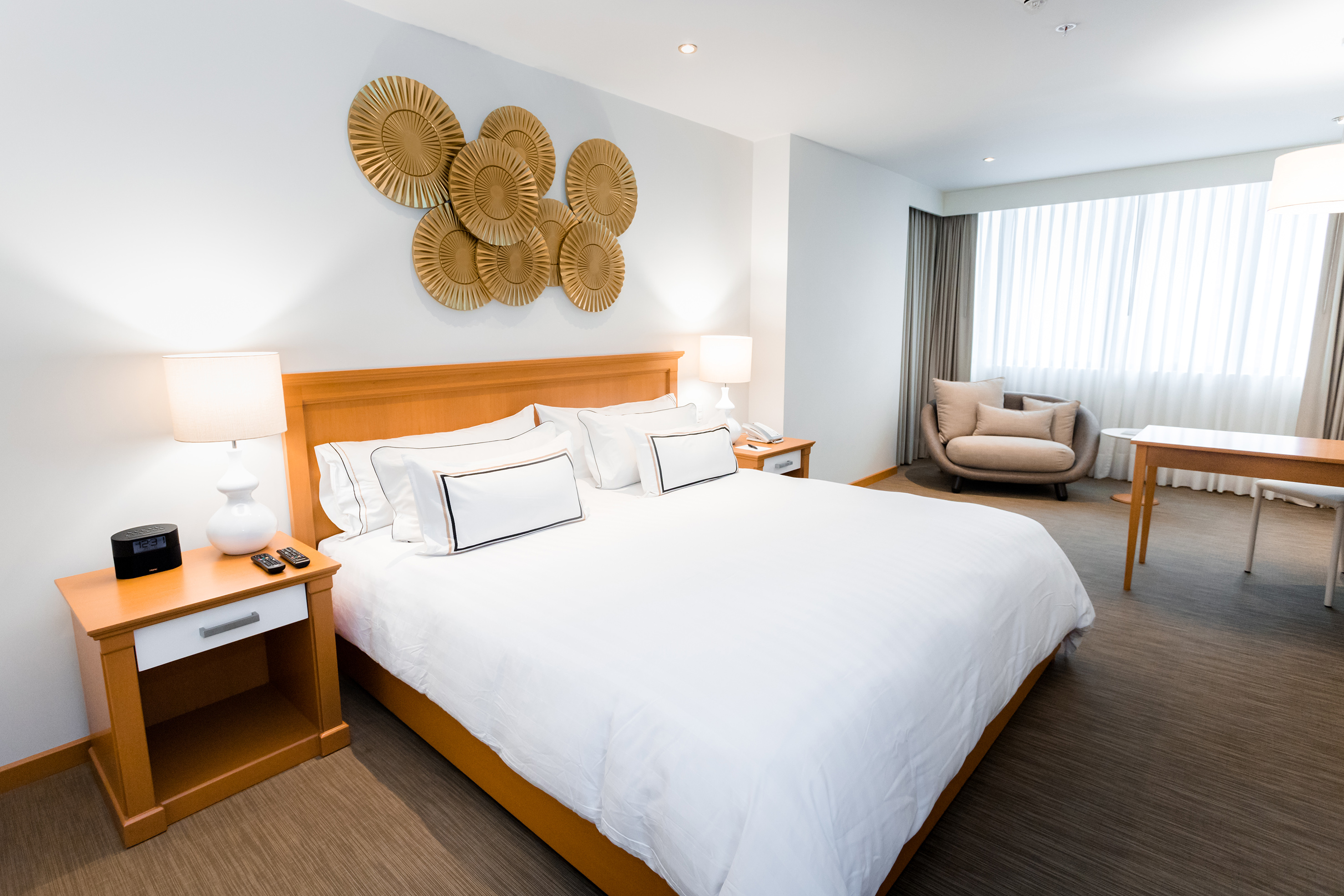 Spacious modern hotel bedroom with king bed, gold decor, armchair, and large window.