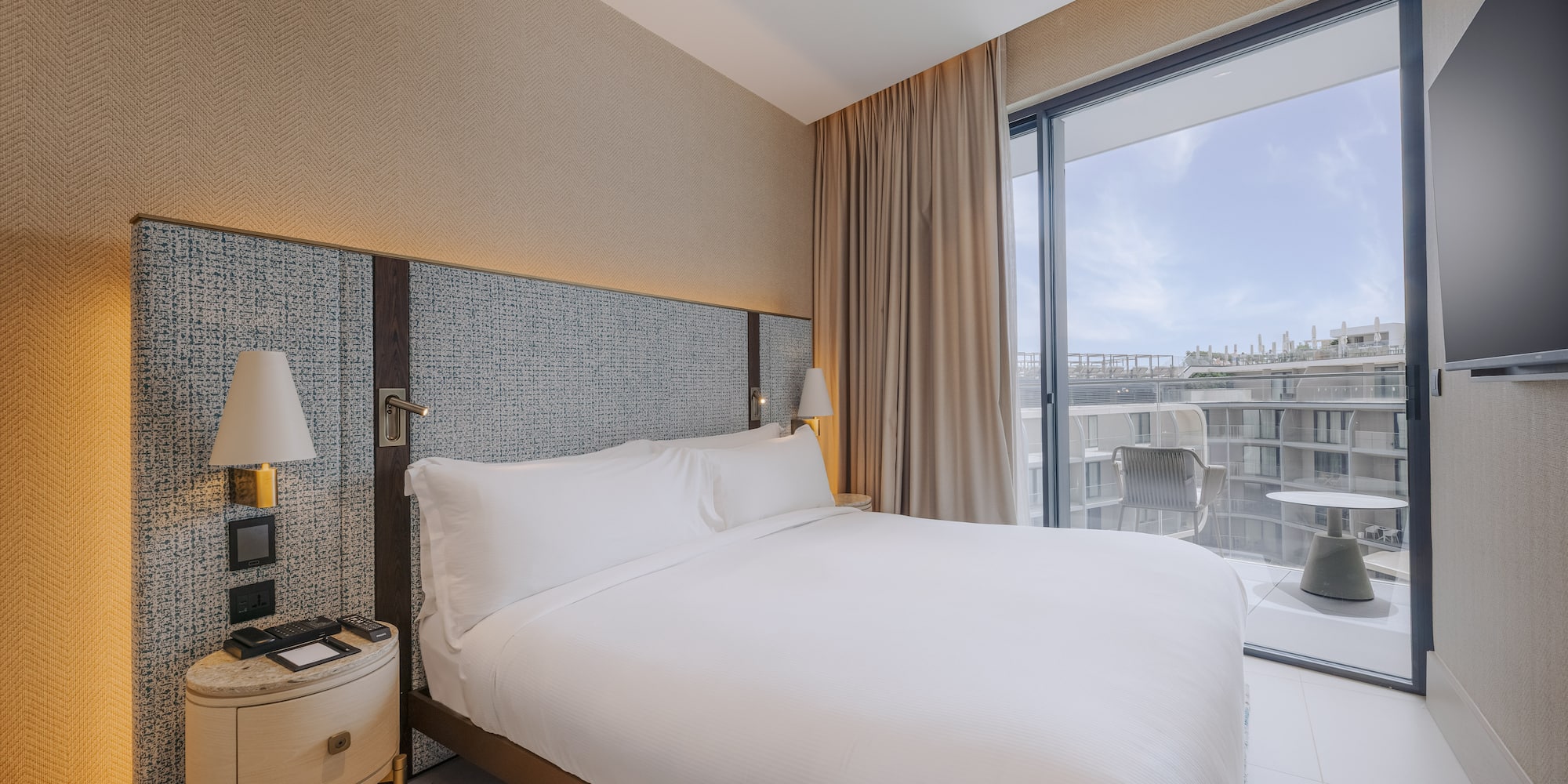 Modern hotel bedroom with large white bed, textured headboard, and balcony.