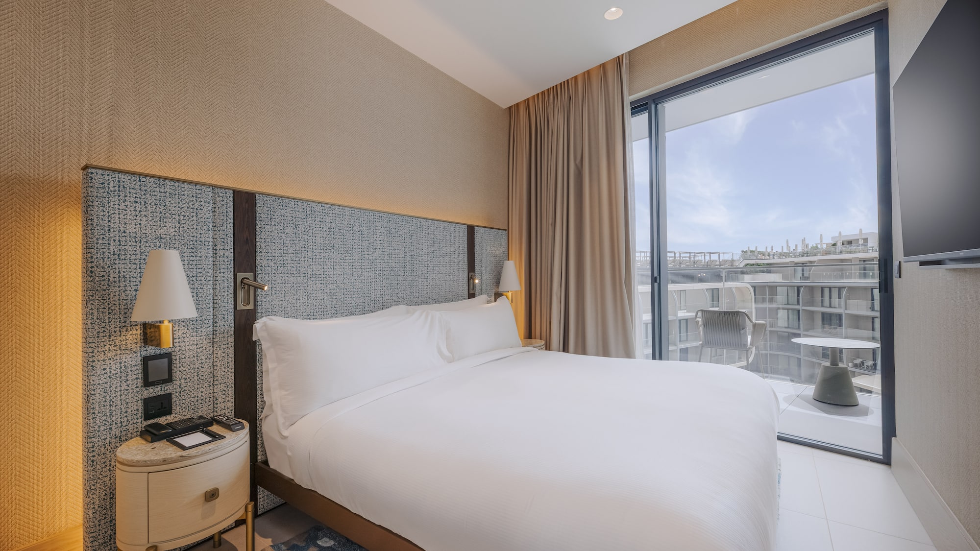 Modern hotel bedroom with large white bed, textured headboard, and balcony.