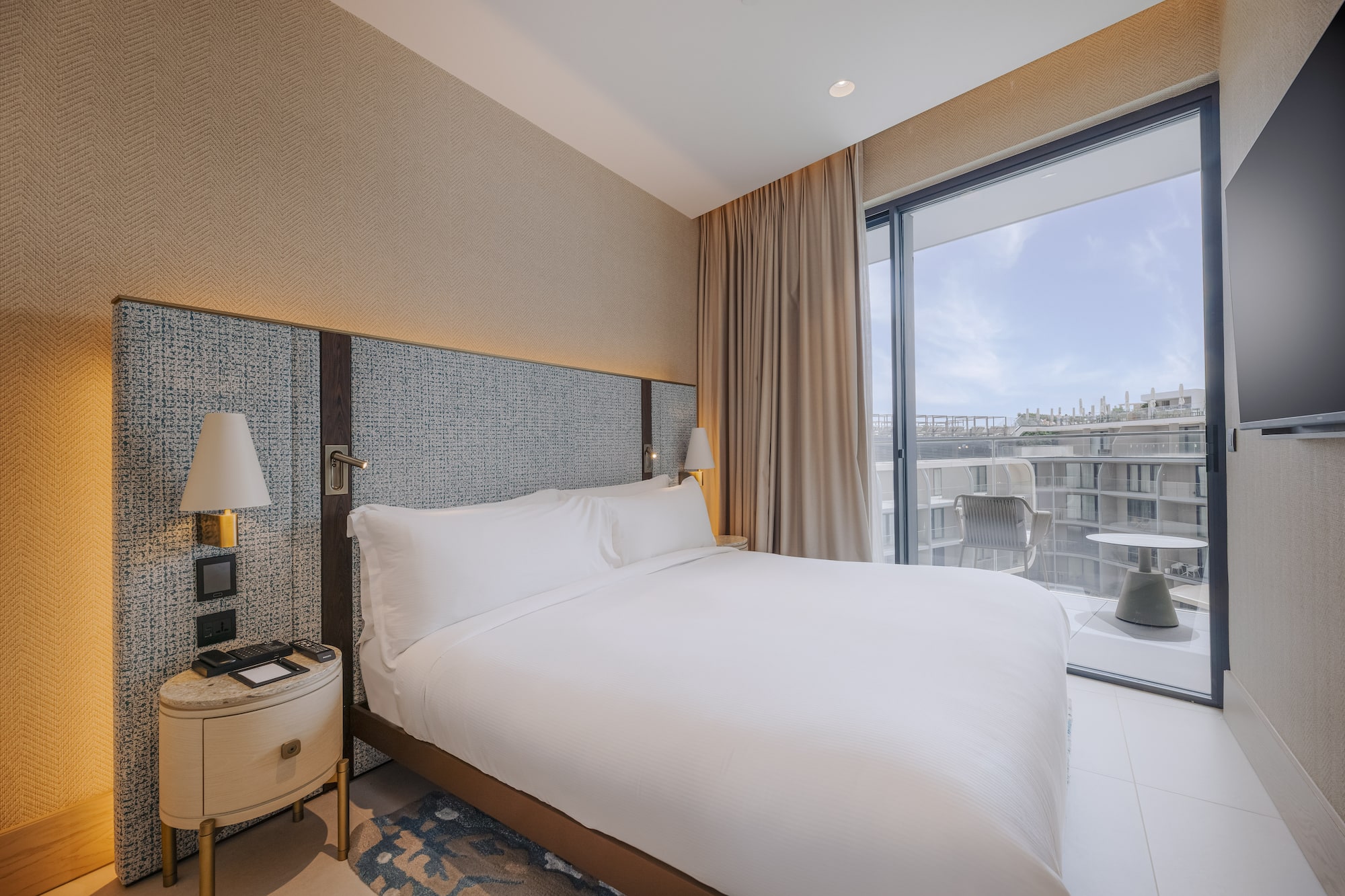Modern hotel bedroom with large white bed, textured headboard, and balcony.