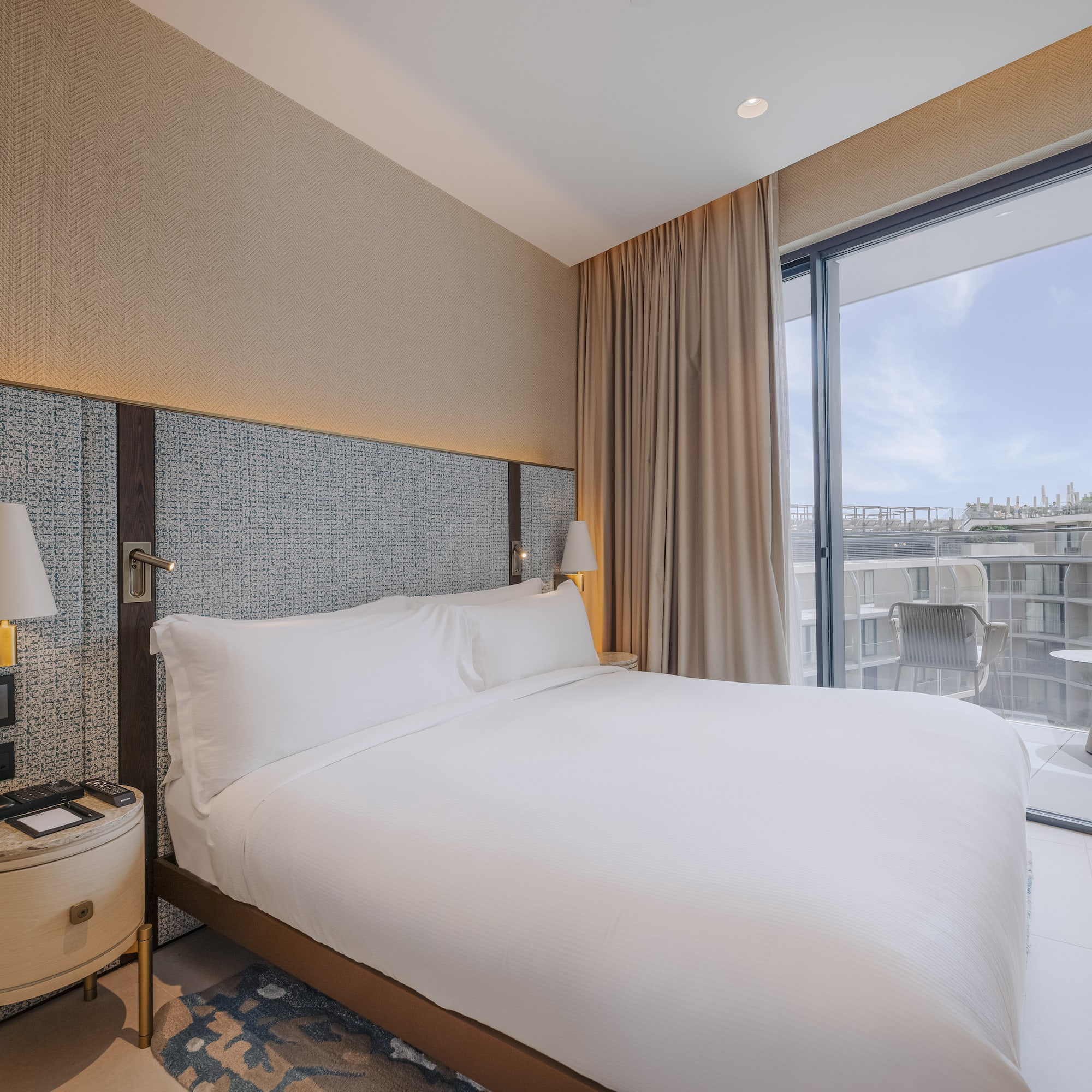 Modern hotel bedroom with large white bed, textured headboard, and balcony.