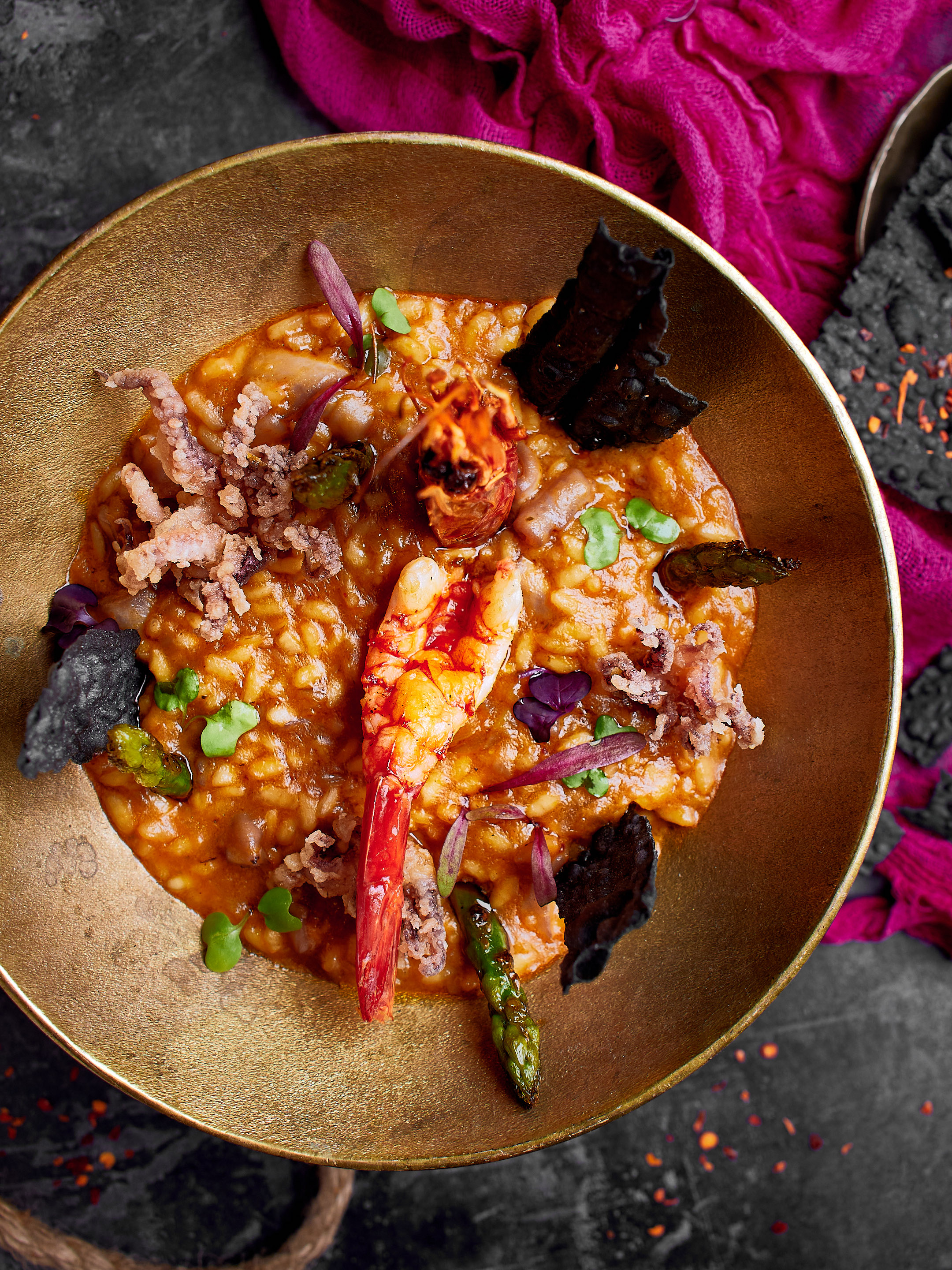 Seafood risotto with shrimp, calamari, asparagus, and black crisps in golden bowl.
