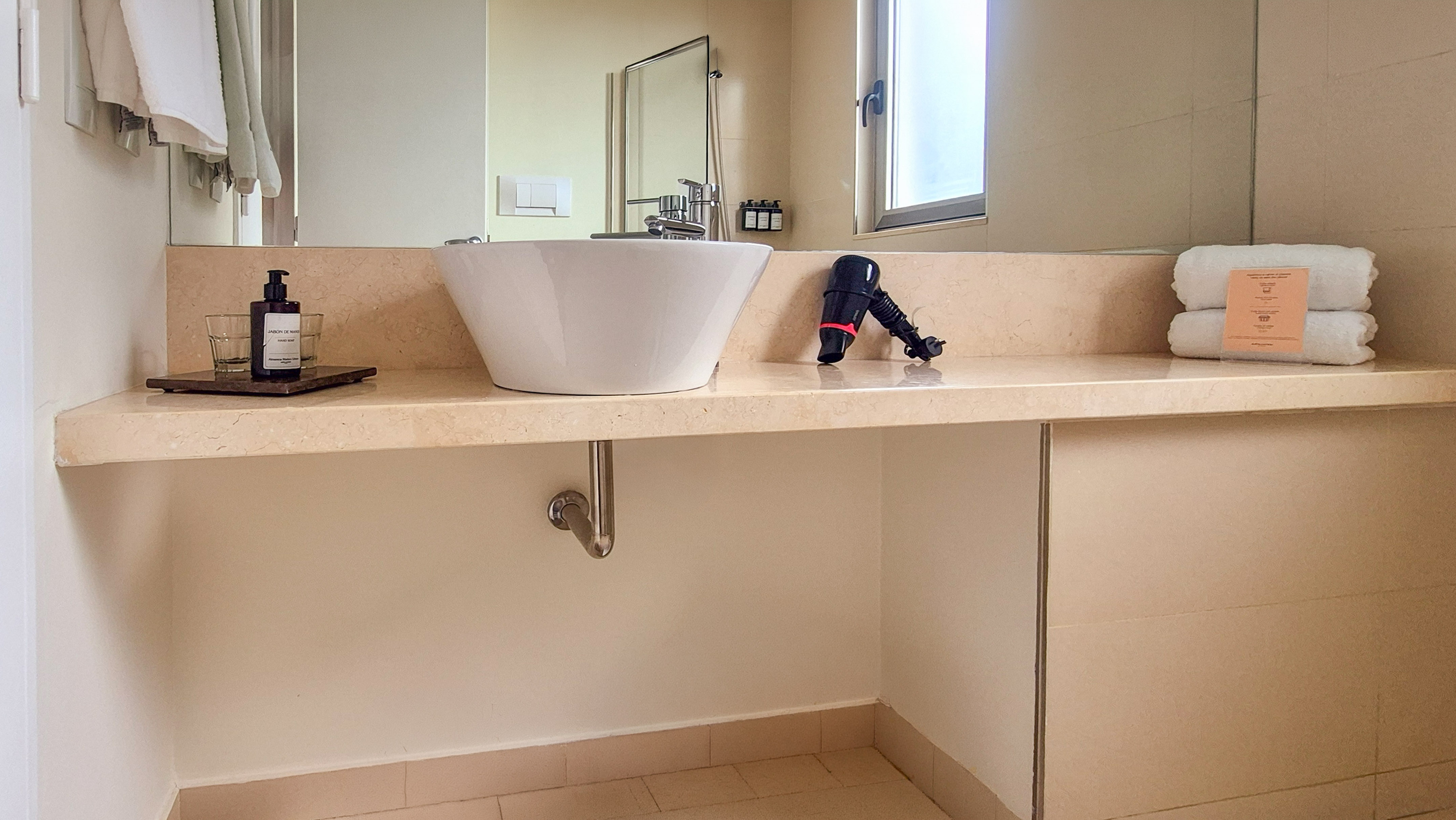 Modern hotel bathroom with vessel sink, beige countertop, hairdryer, and towels.