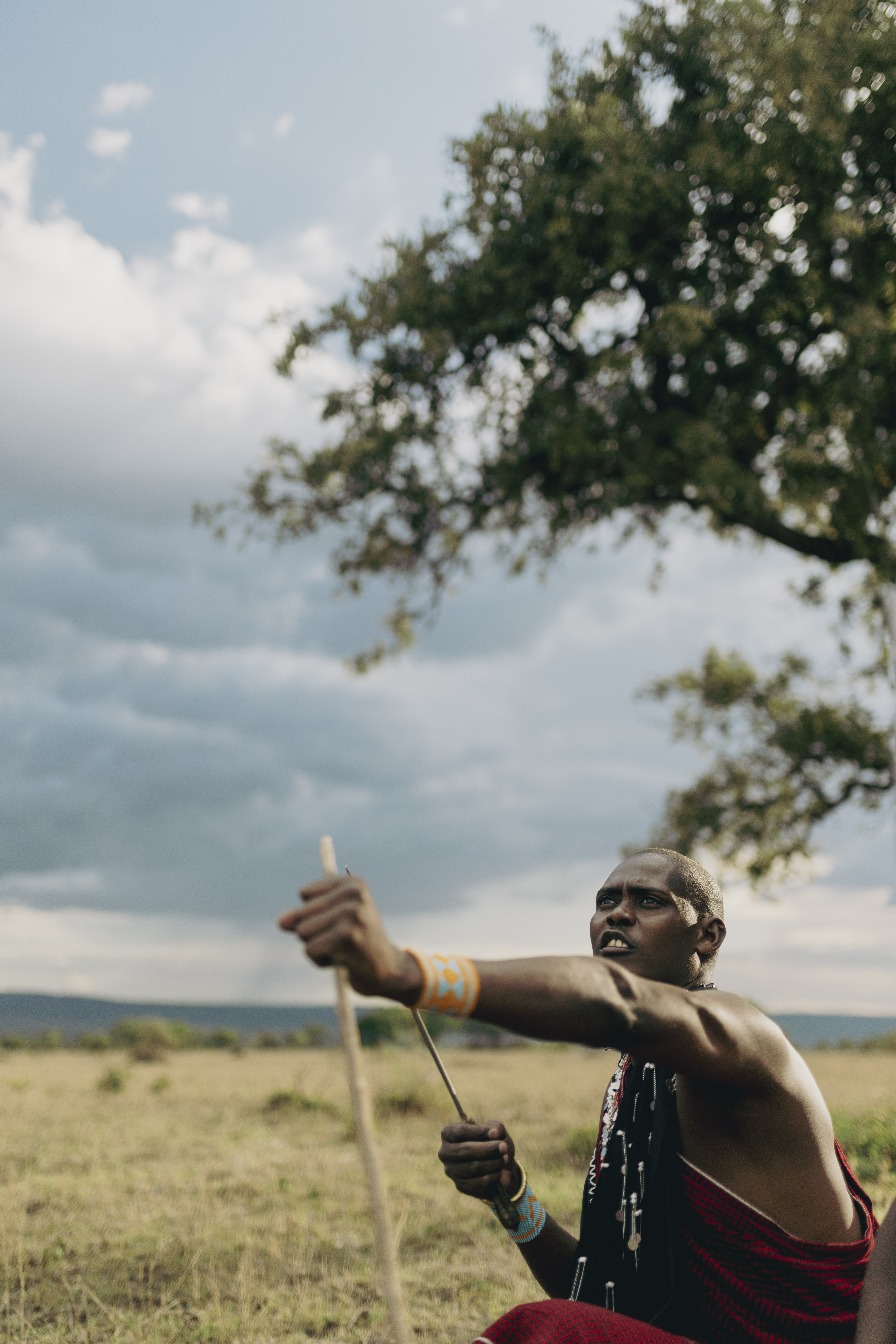 Maasai warrior in traditional dress aiming a spear in the savanna landscape.