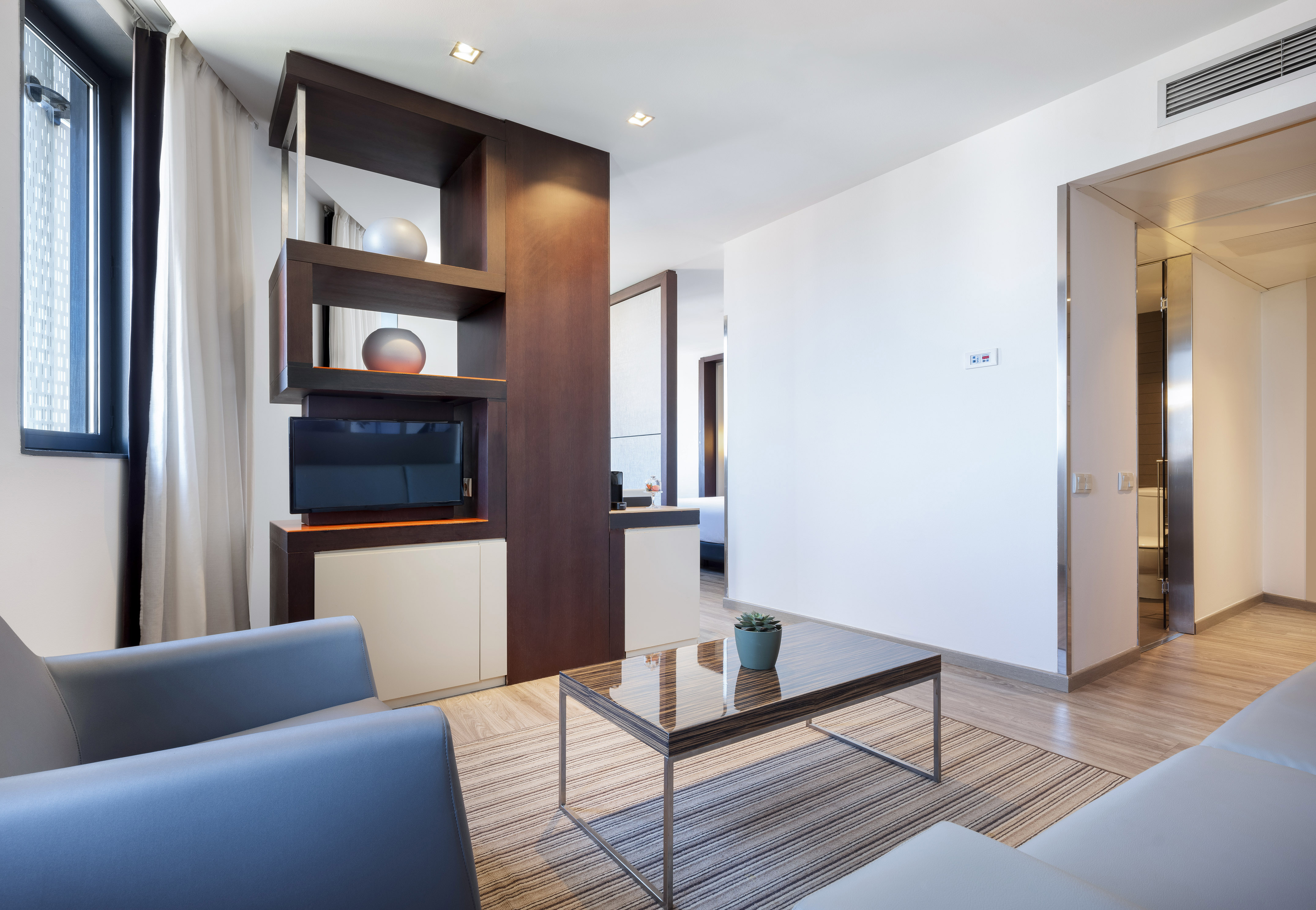 Modern hotel suite living room with grey sofa, wood coffee table, and TV unit.