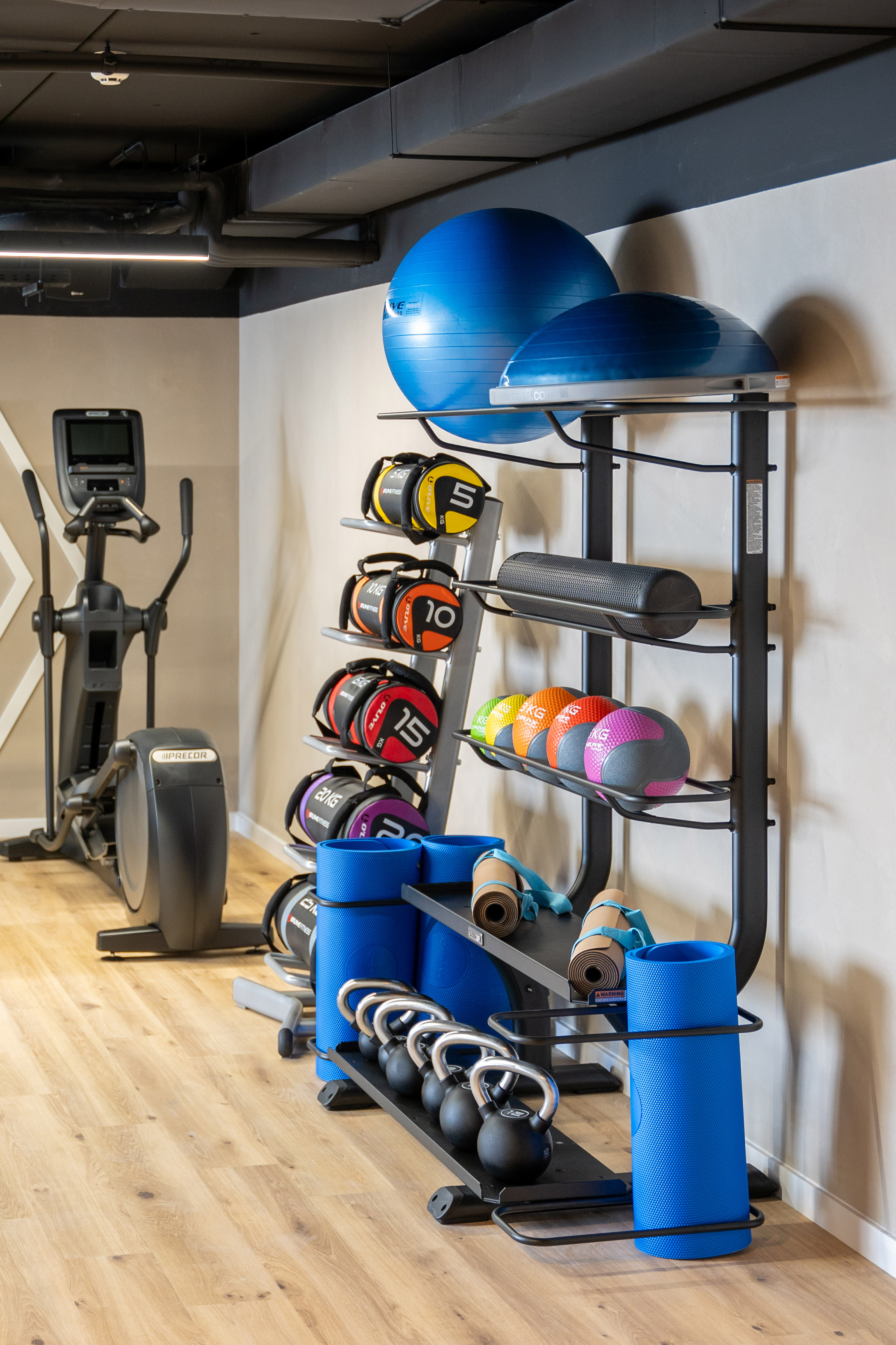 Gym with elliptical, weights, exercise balls, yoga mats, foam roller on wood floor.