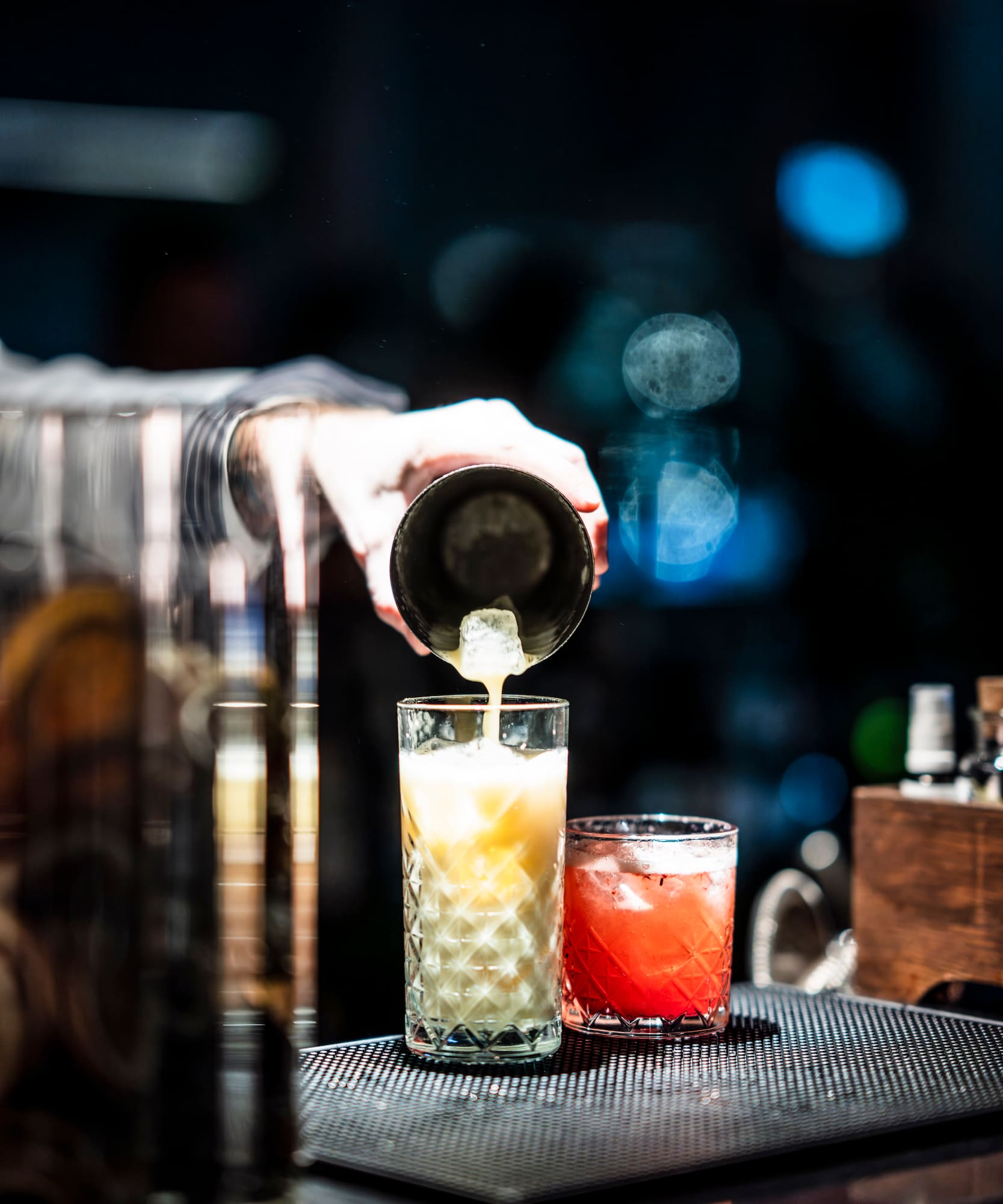 Bartender pours creamy cocktail from shaker into glass; red drink nearby on bar.
