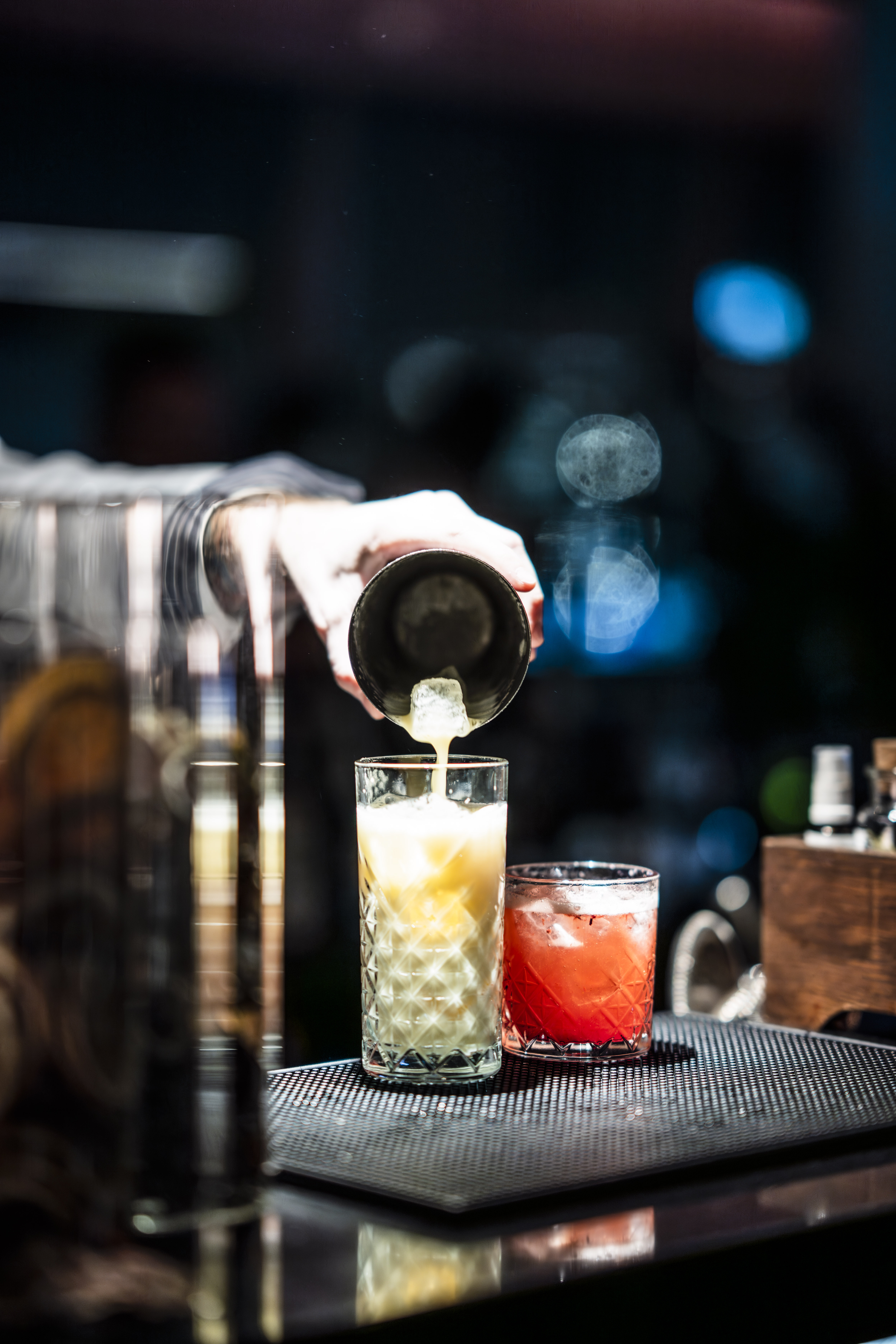 Bartender pours creamy cocktail from shaker into glass; red drink nearby on bar.