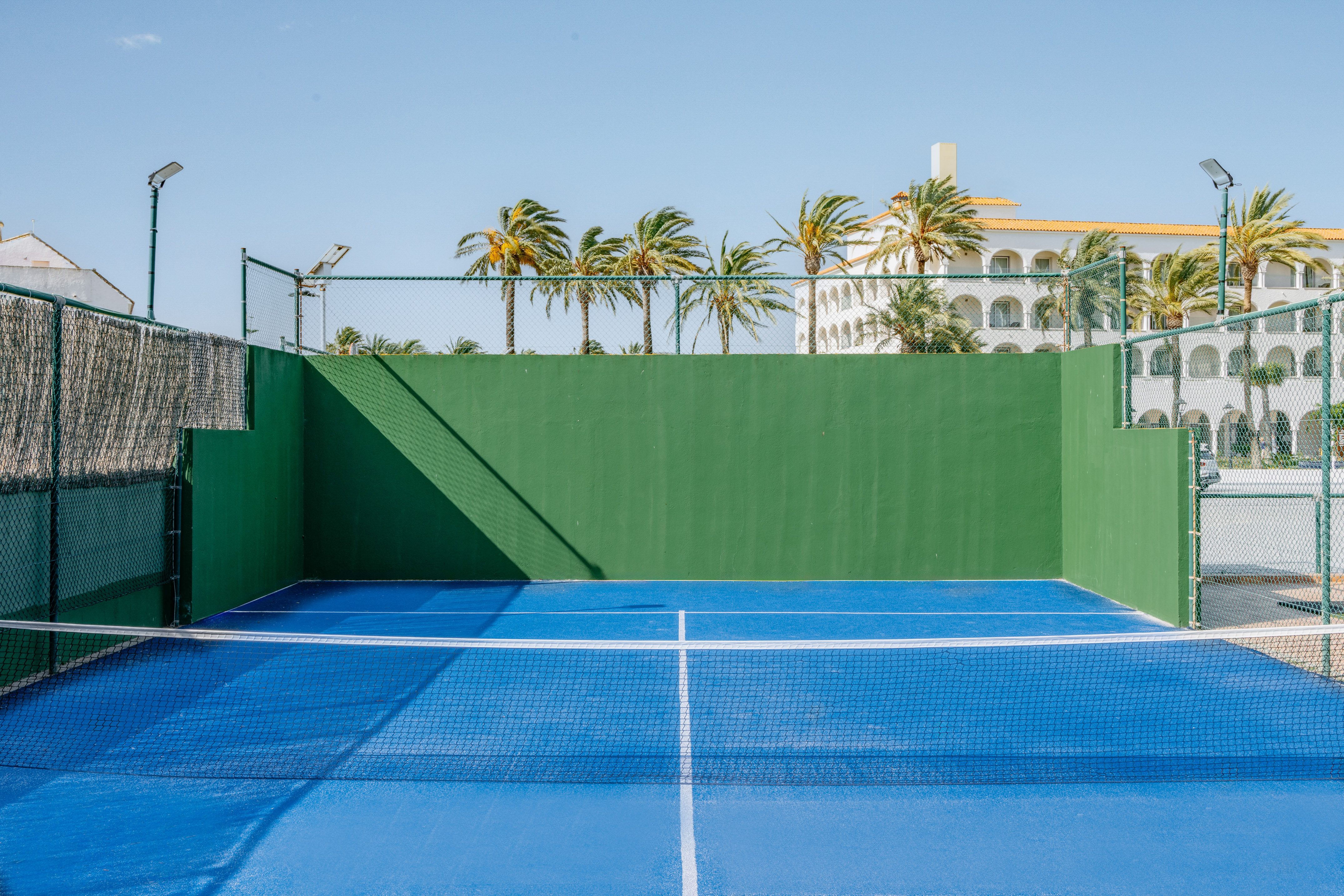Blue padel court with green walls, net, palm trees, and resort.