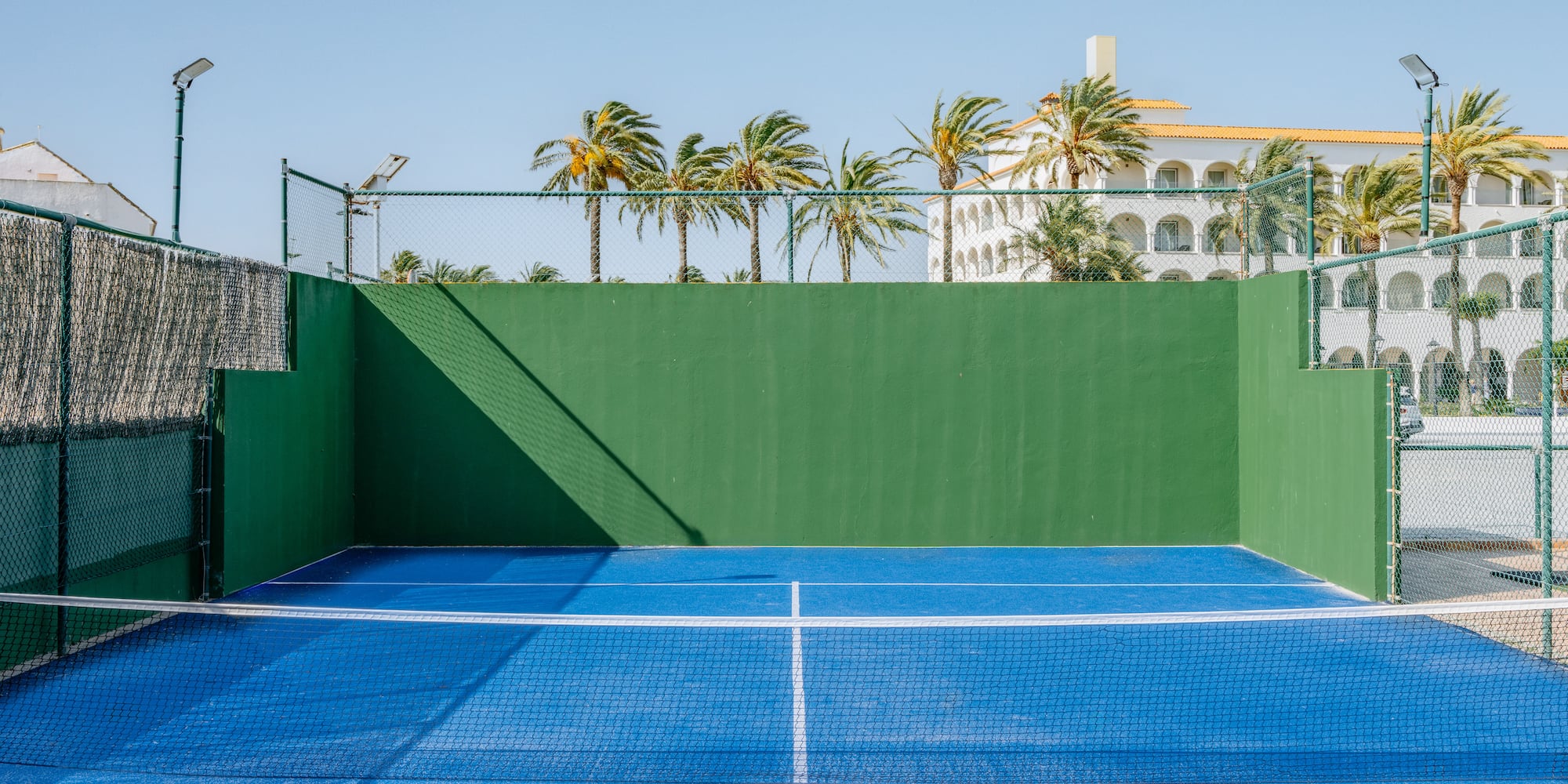 Blue padel court with green walls, net, palm trees, and resort.