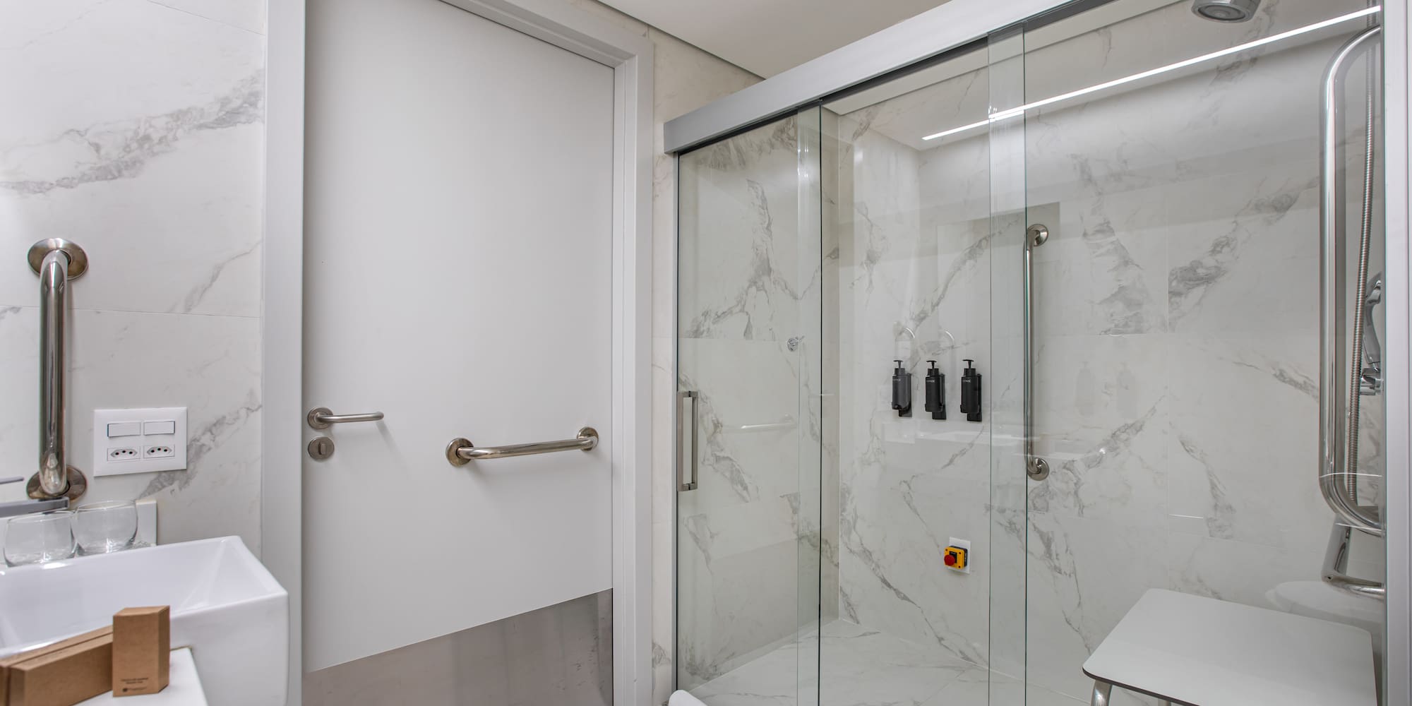 Accessible bathroom with grab bars, shower seat, marble tiles, and glass enclosure.