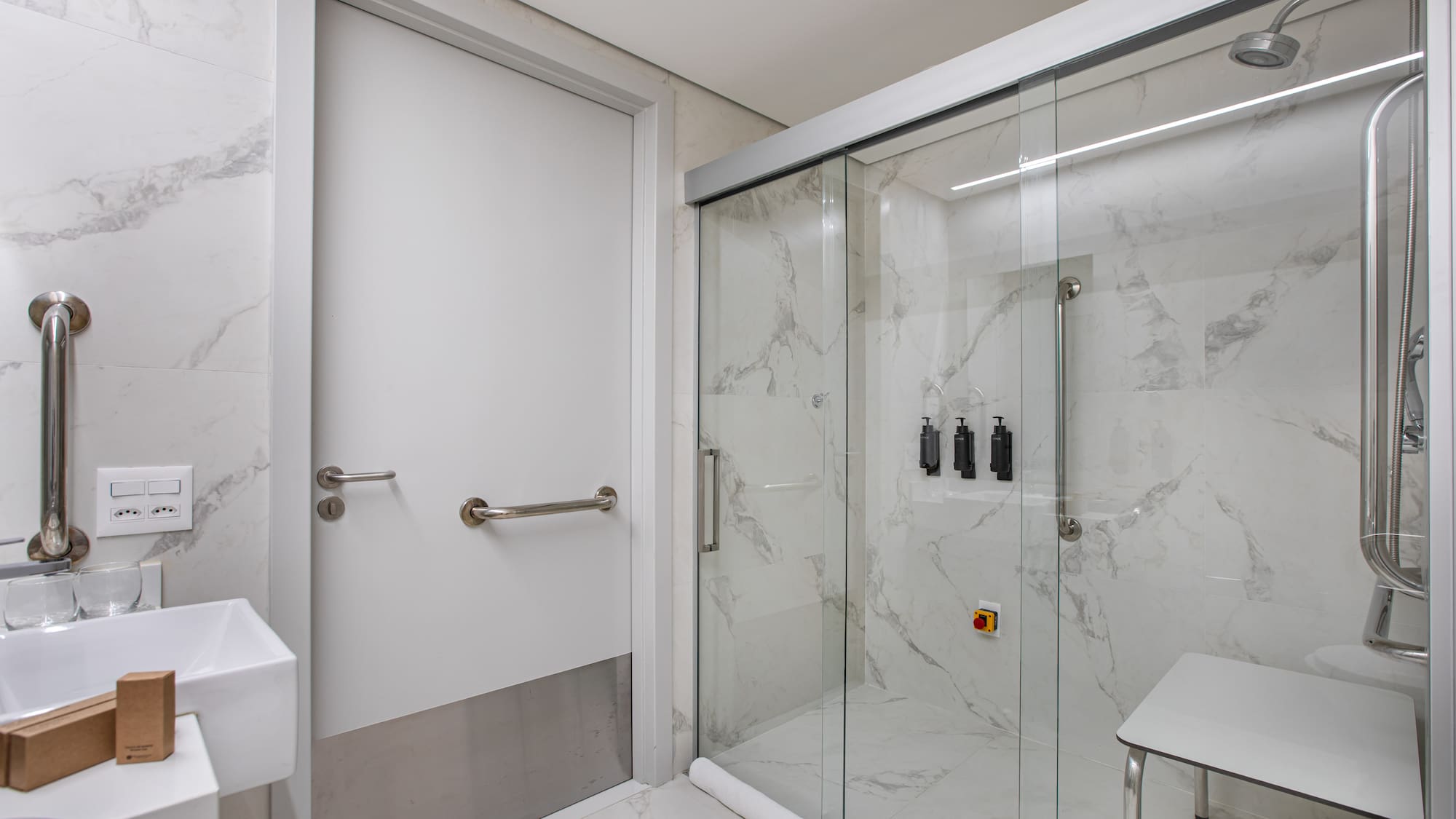 Accessible bathroom with grab bars, shower seat, marble tiles, and glass enclosure.
