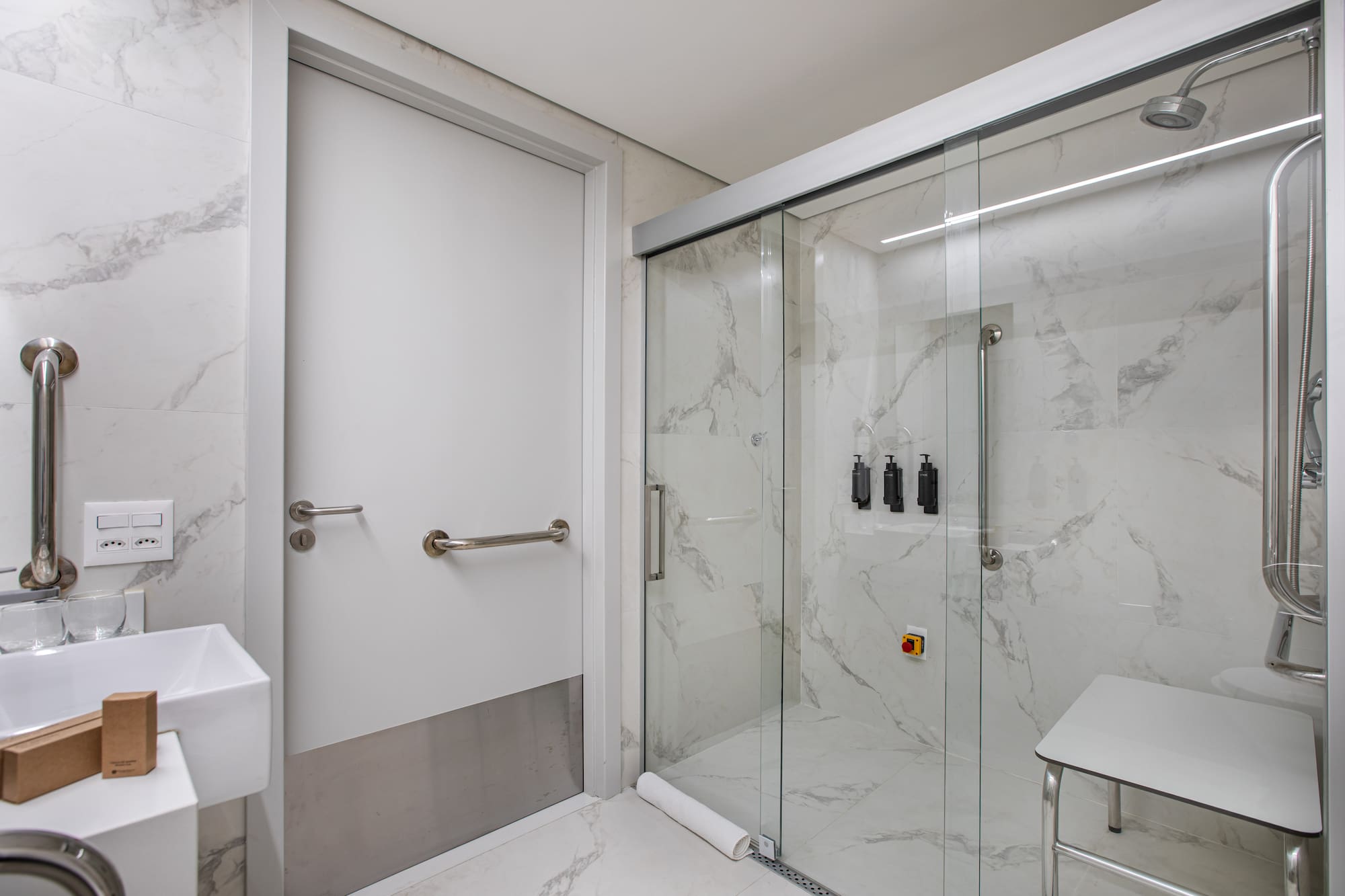 Accessible bathroom with grab bars, shower seat, marble tiles, and glass enclosure.