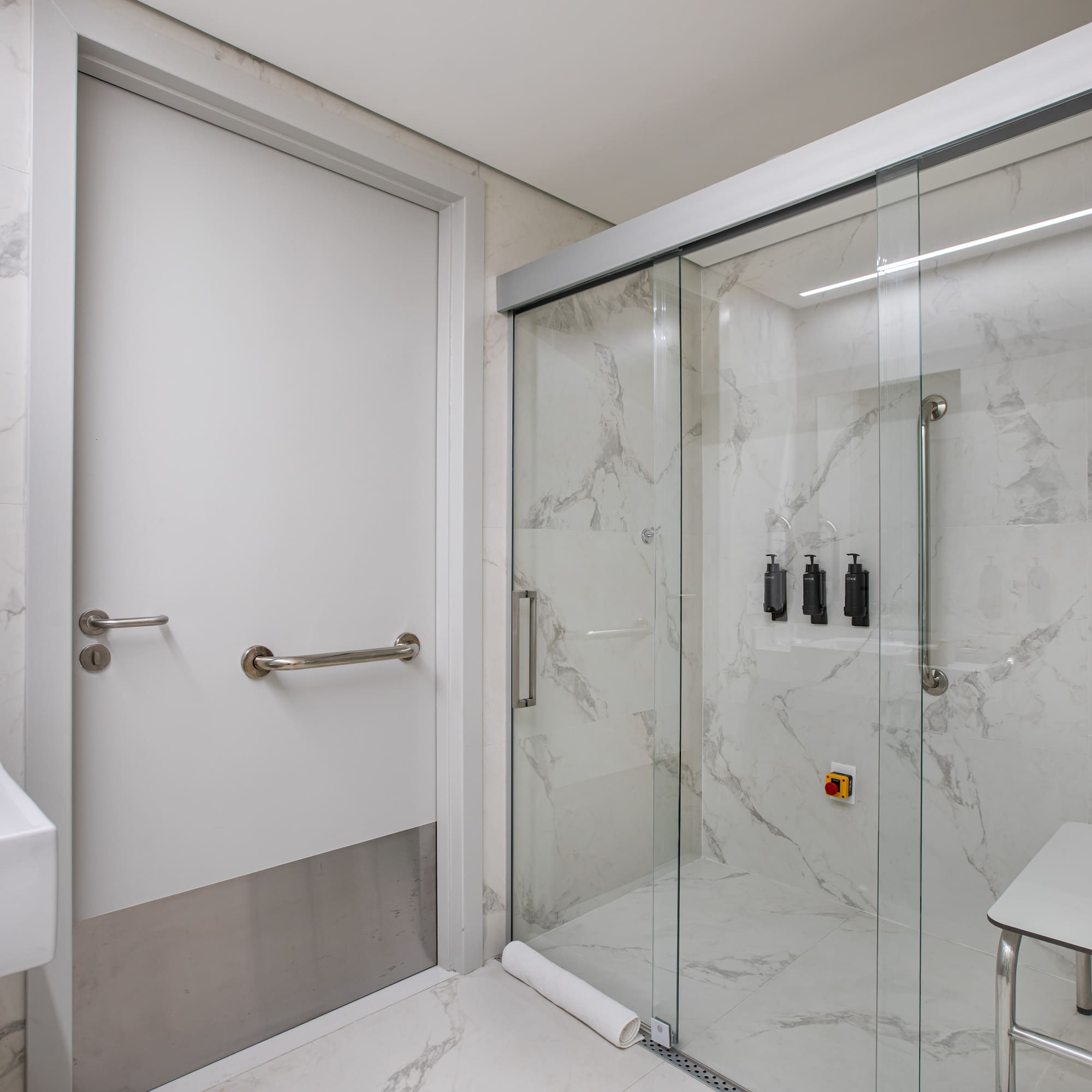 Accessible bathroom with grab bars, shower seat, marble tiles, and glass enclosure.