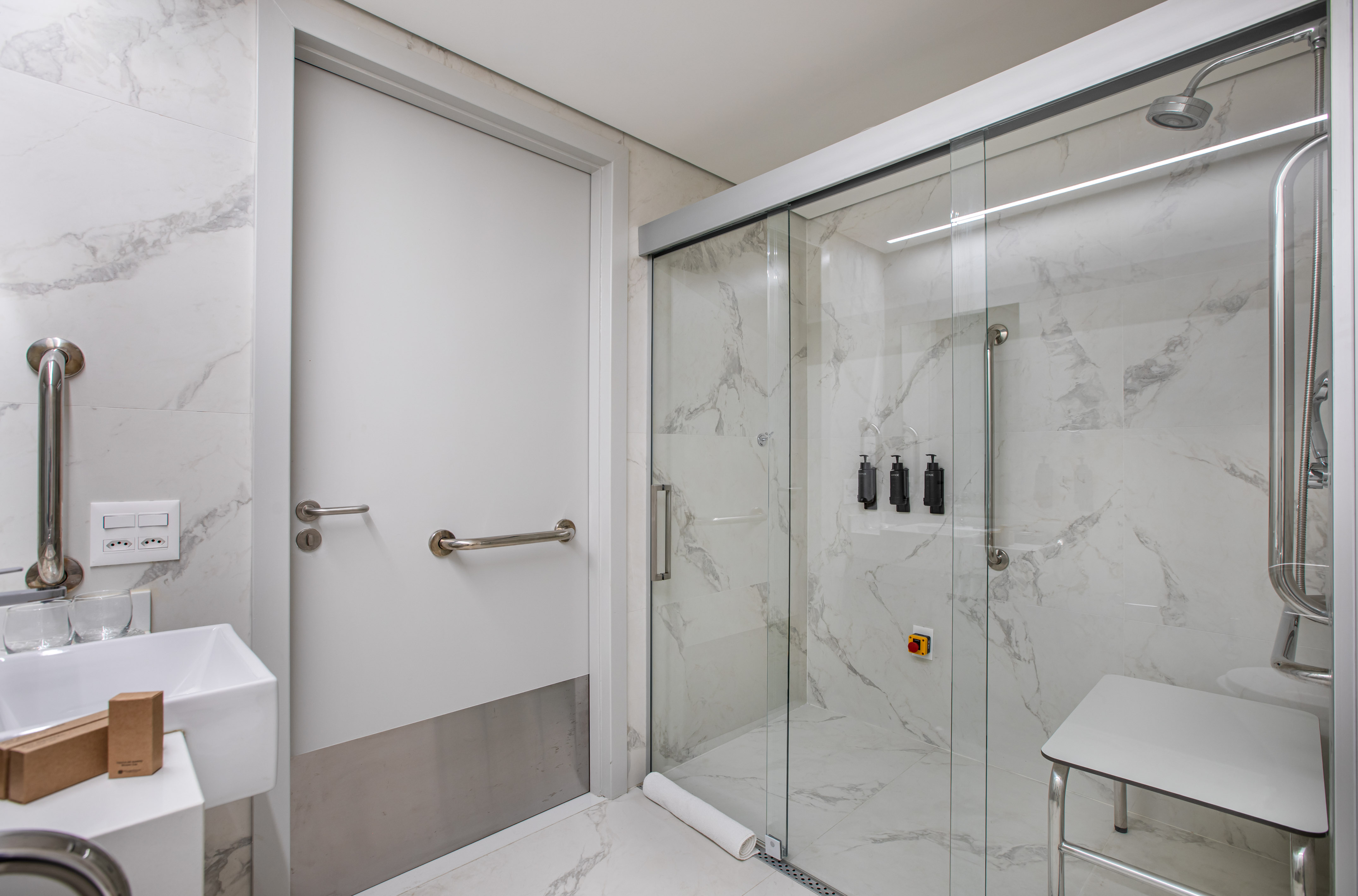 Accessible bathroom with grab bars, shower seat, marble tiles, and glass enclosure.