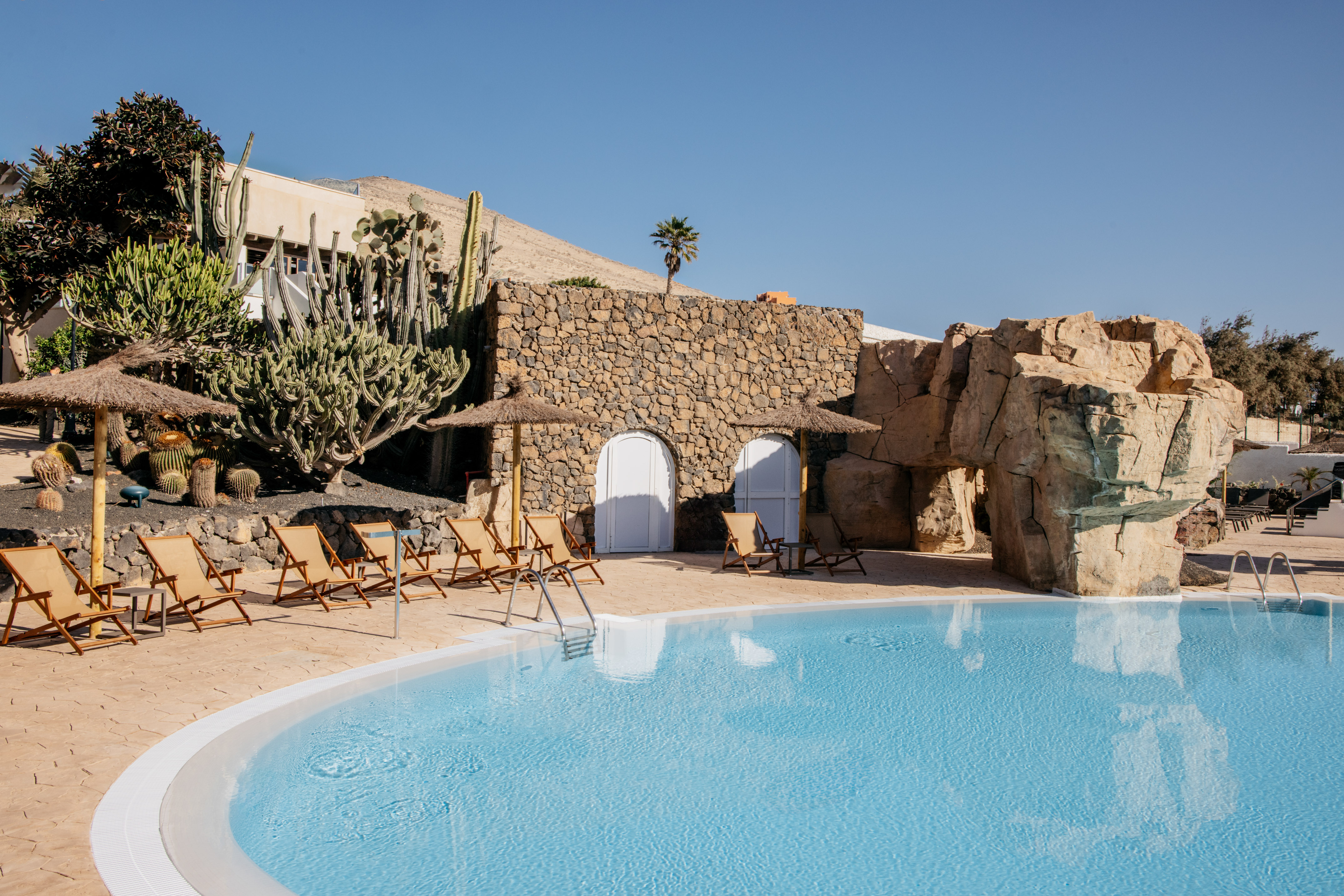 Hotel pool area with sun loungers, cacti, stone building, and desert.
