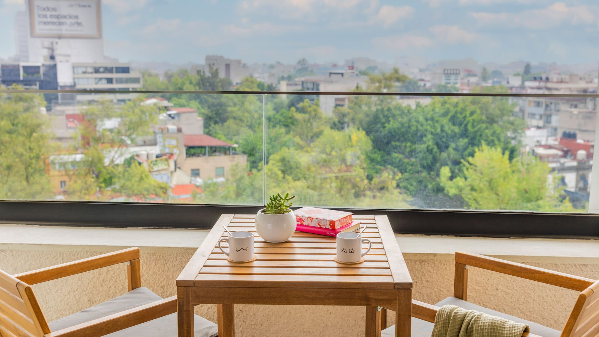 Balcón moderno con mesa de madera, dos sillas y vista urbana verde.