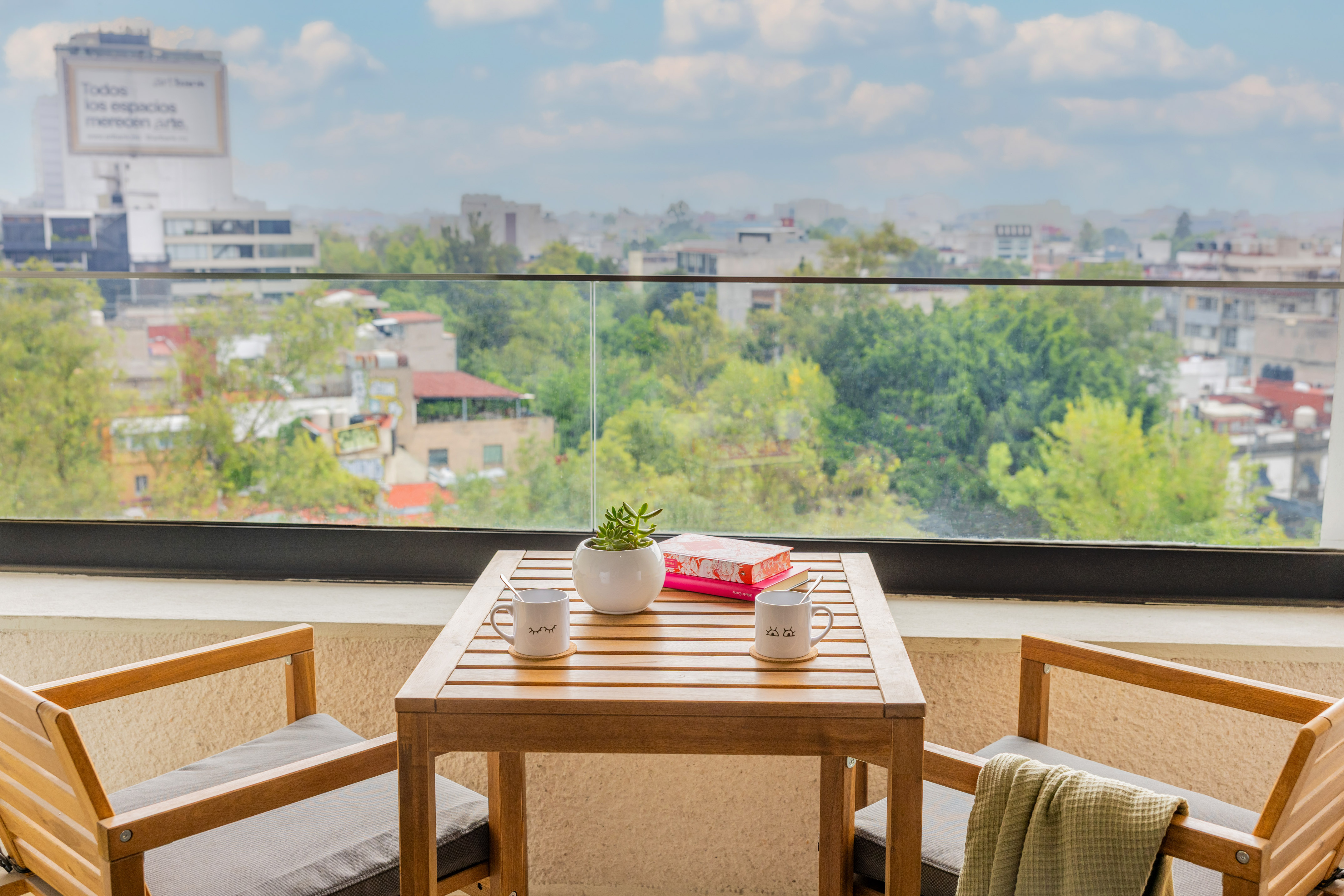 Apartment balcony with wooden table, two chairs, coffee, plant, and city view.
