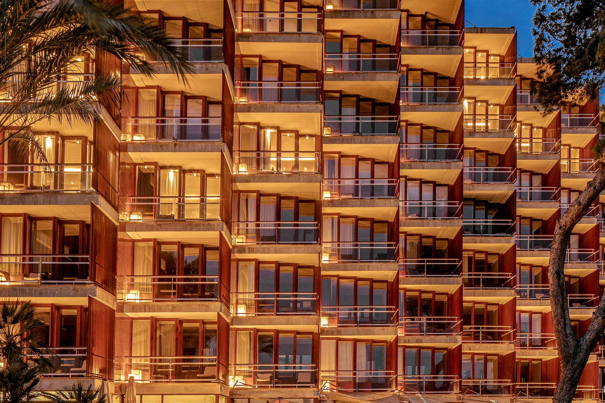 Luxury hotel exterior with lit balconies, reflective pool, and palm trees at twilight.