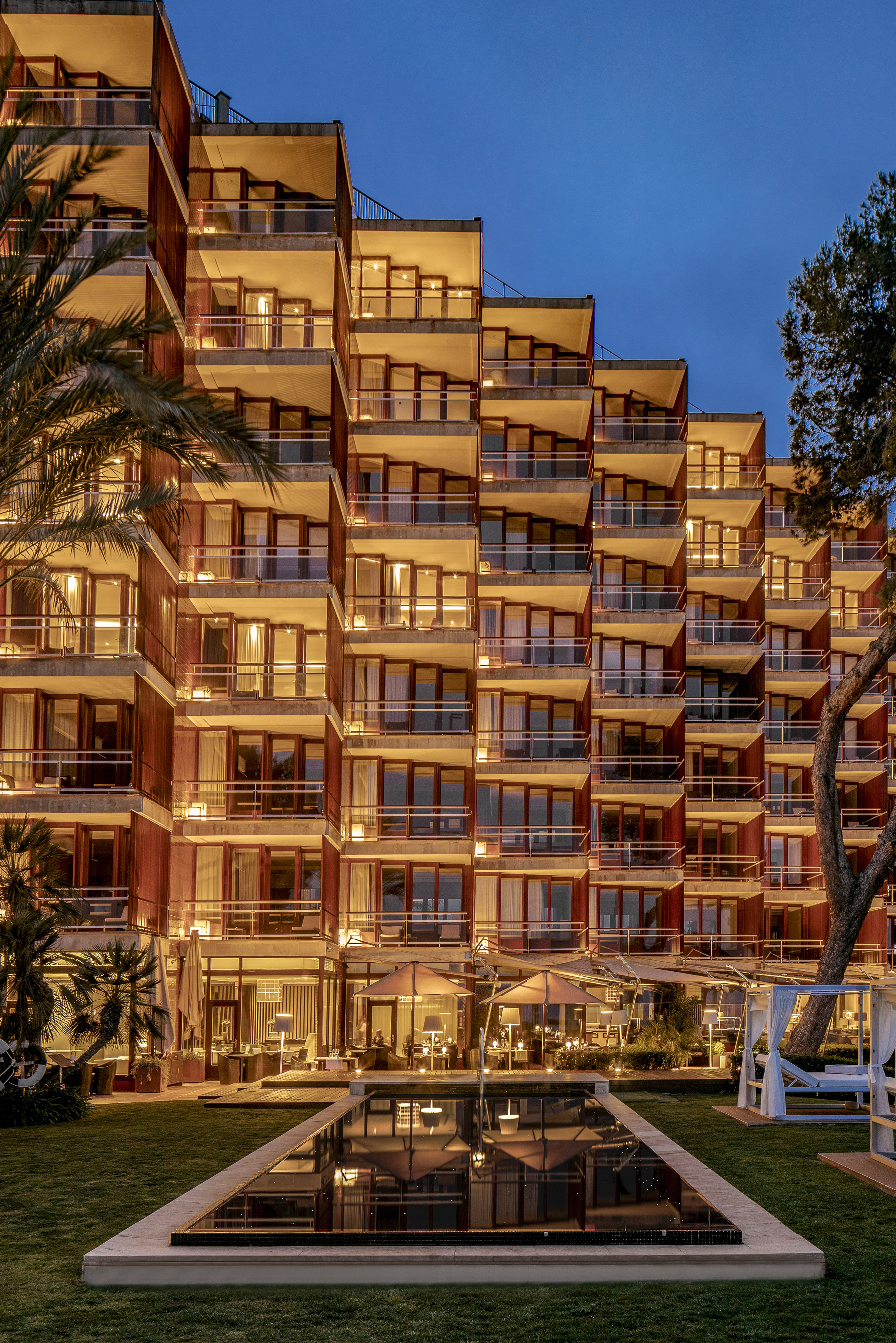 Luxury hotel exterior with lit balconies, reflective pool, and palm trees at twilight.