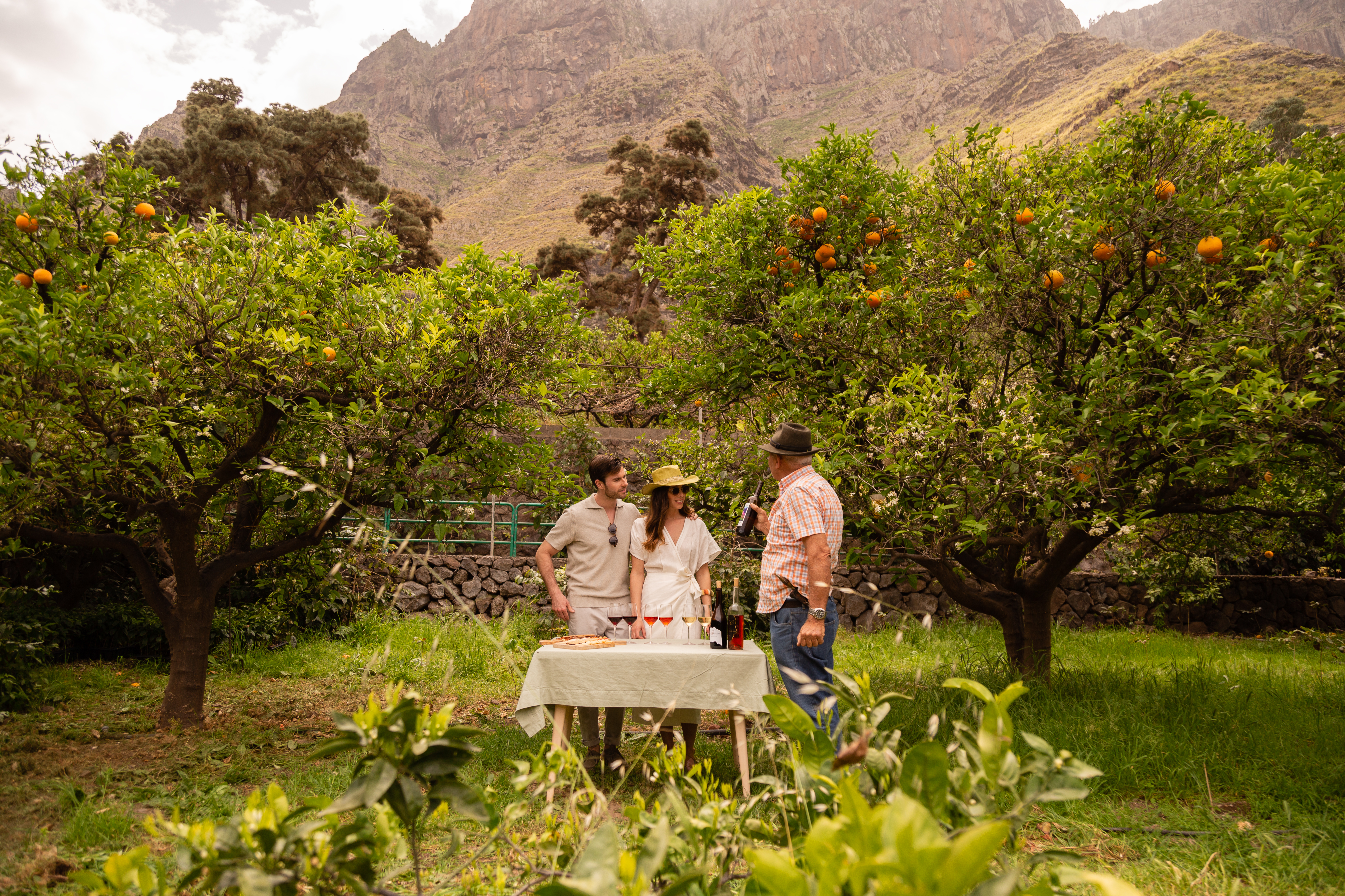 Orange grove wine tasting with couple and man, mountain backdrop.