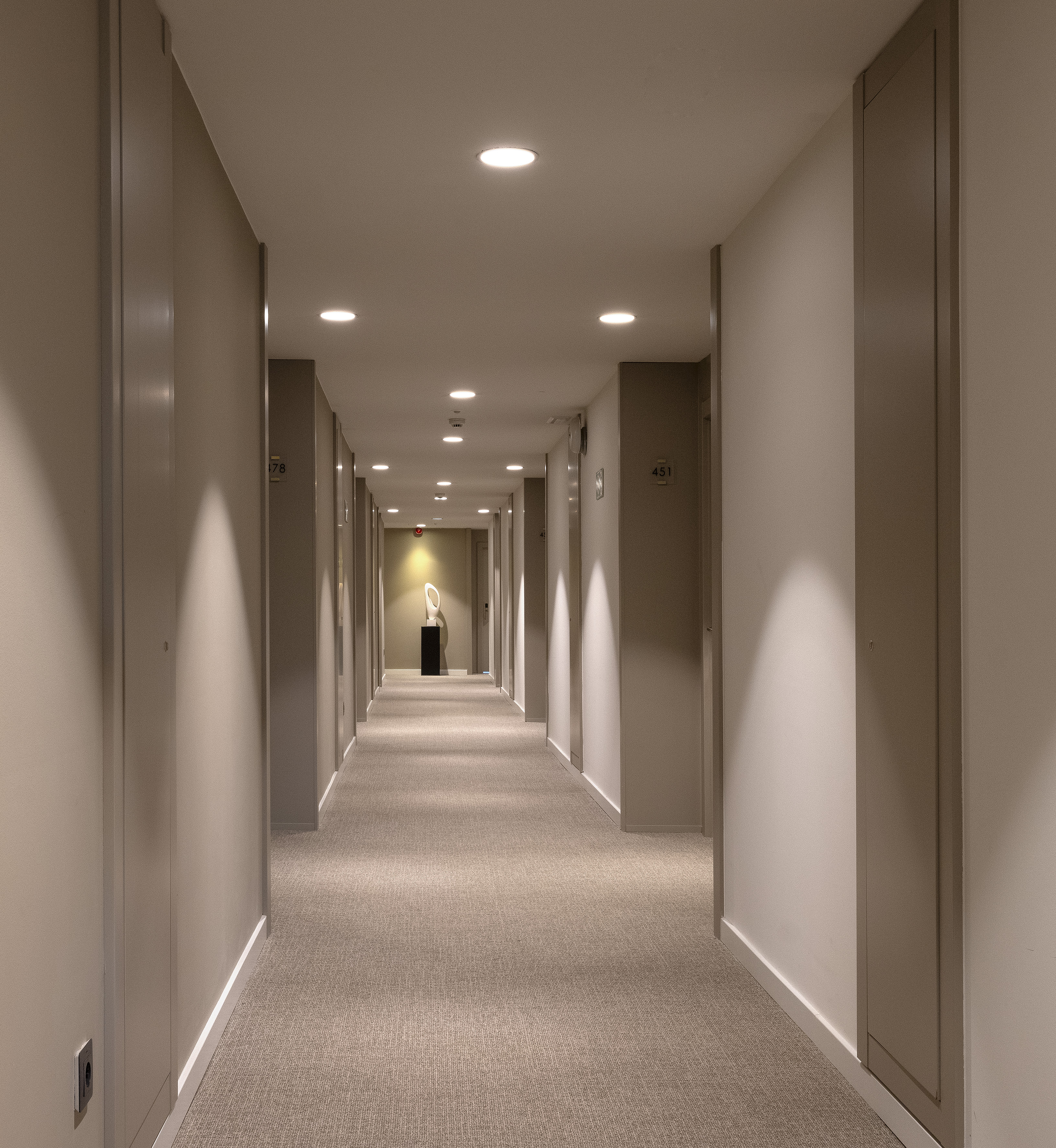 Illuminated hotel corridor with beige walls, patterned carpet, and abstract sculpture.