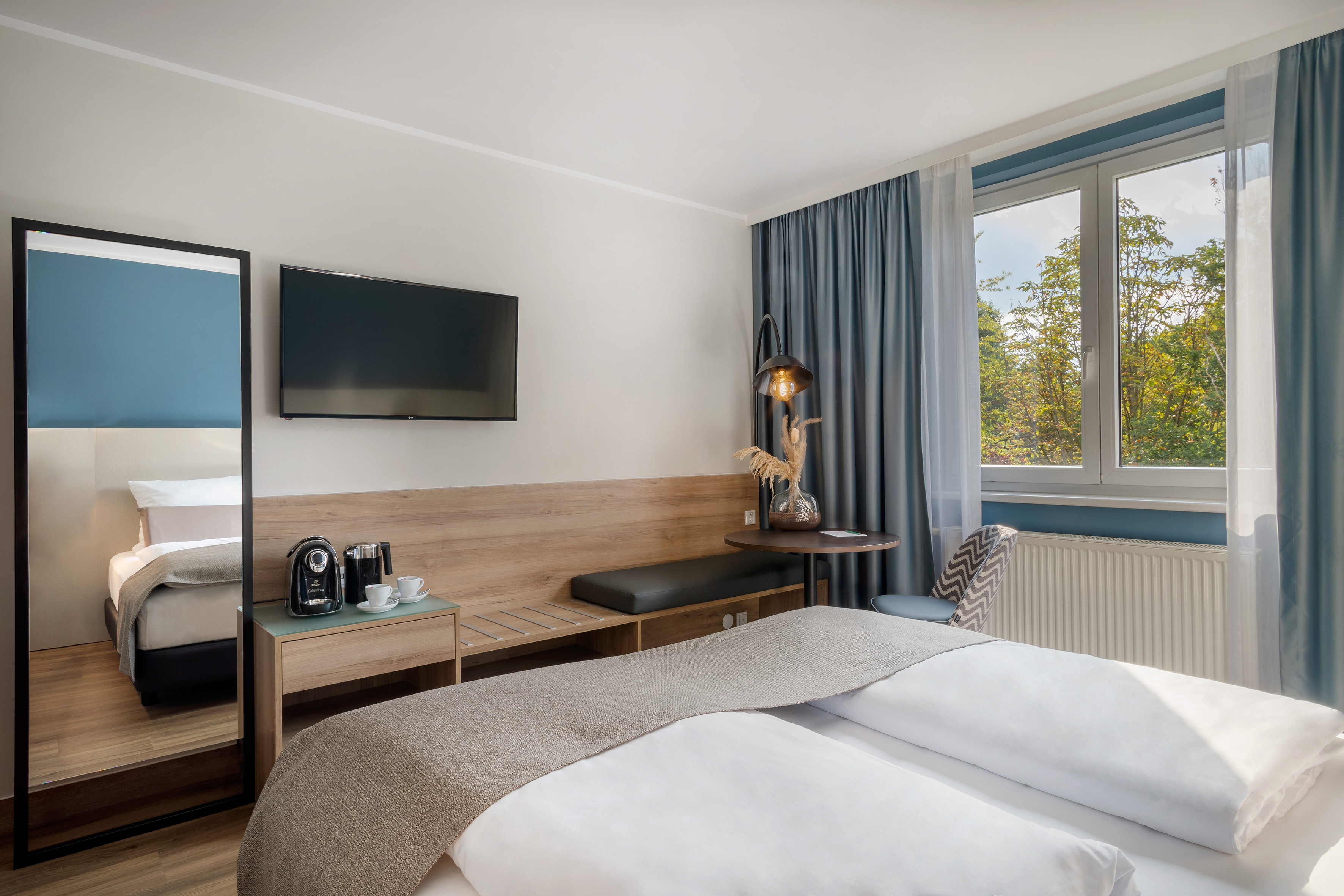 Modern hotel room with bed, TV, coffee station, mirror, and window.
