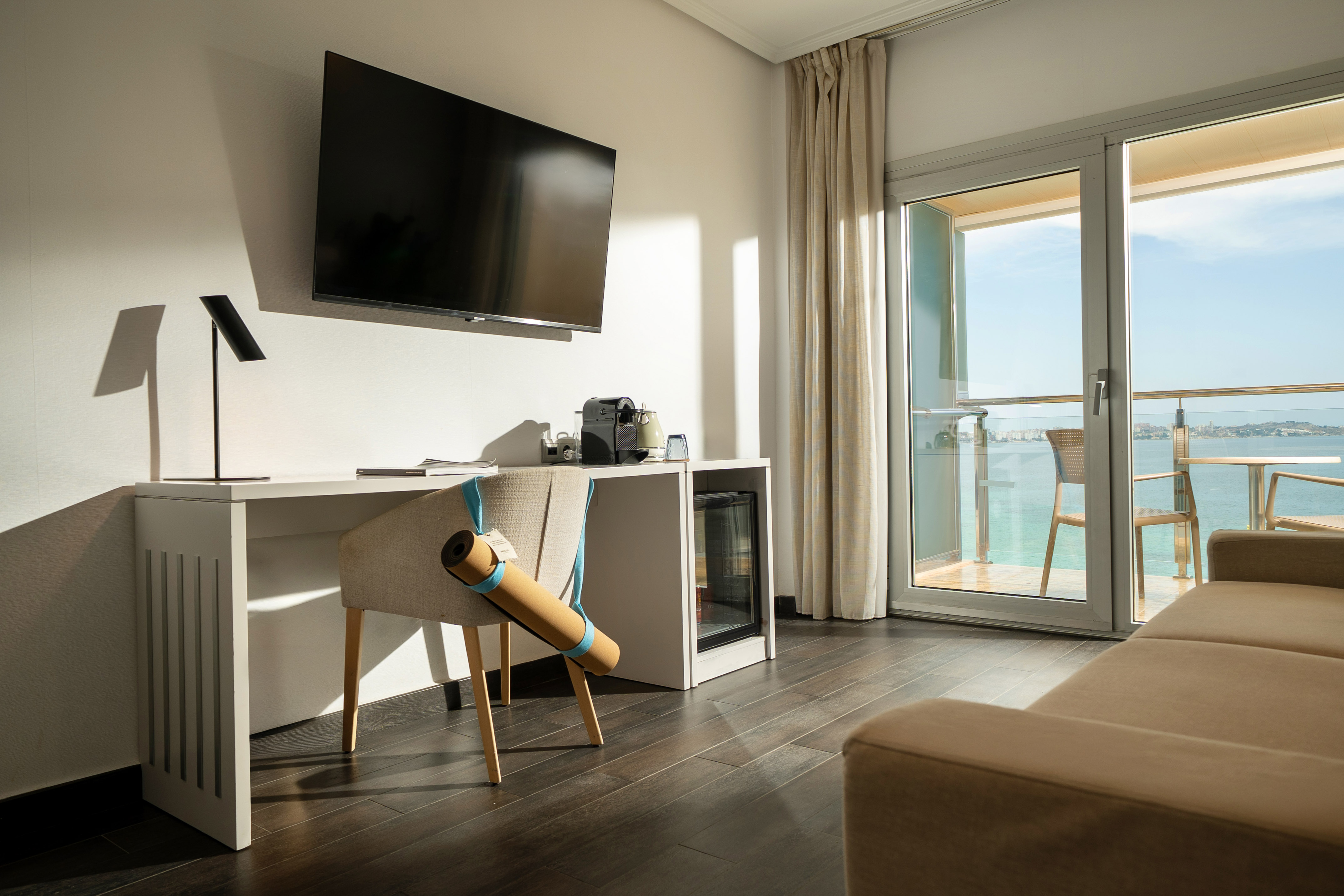 Modern hotel room with sea view balcony, desk, TV, coffee machine, and yoga mat.