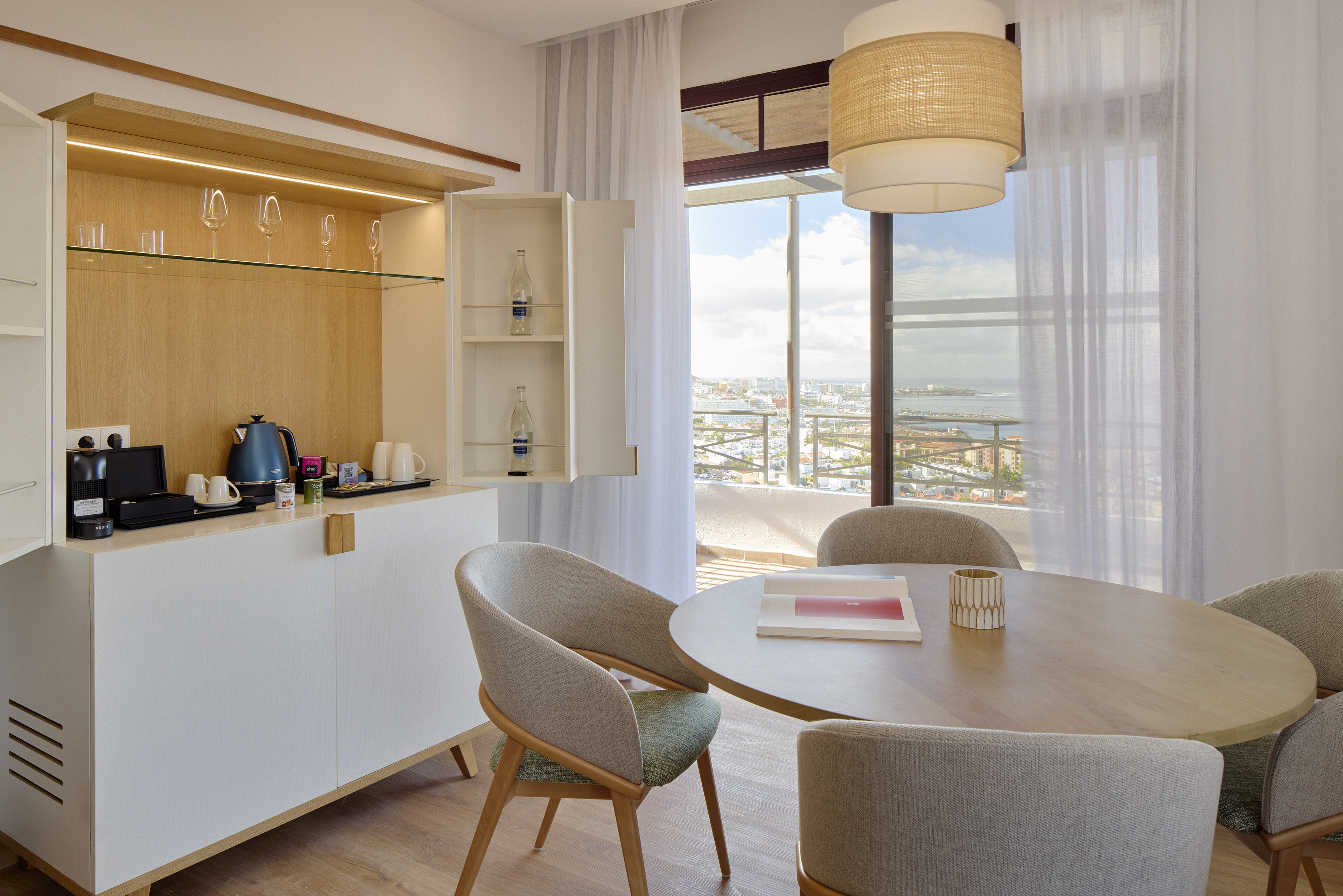 Hotel suite dining area with minibar, coffee machine, and coastal city view.
