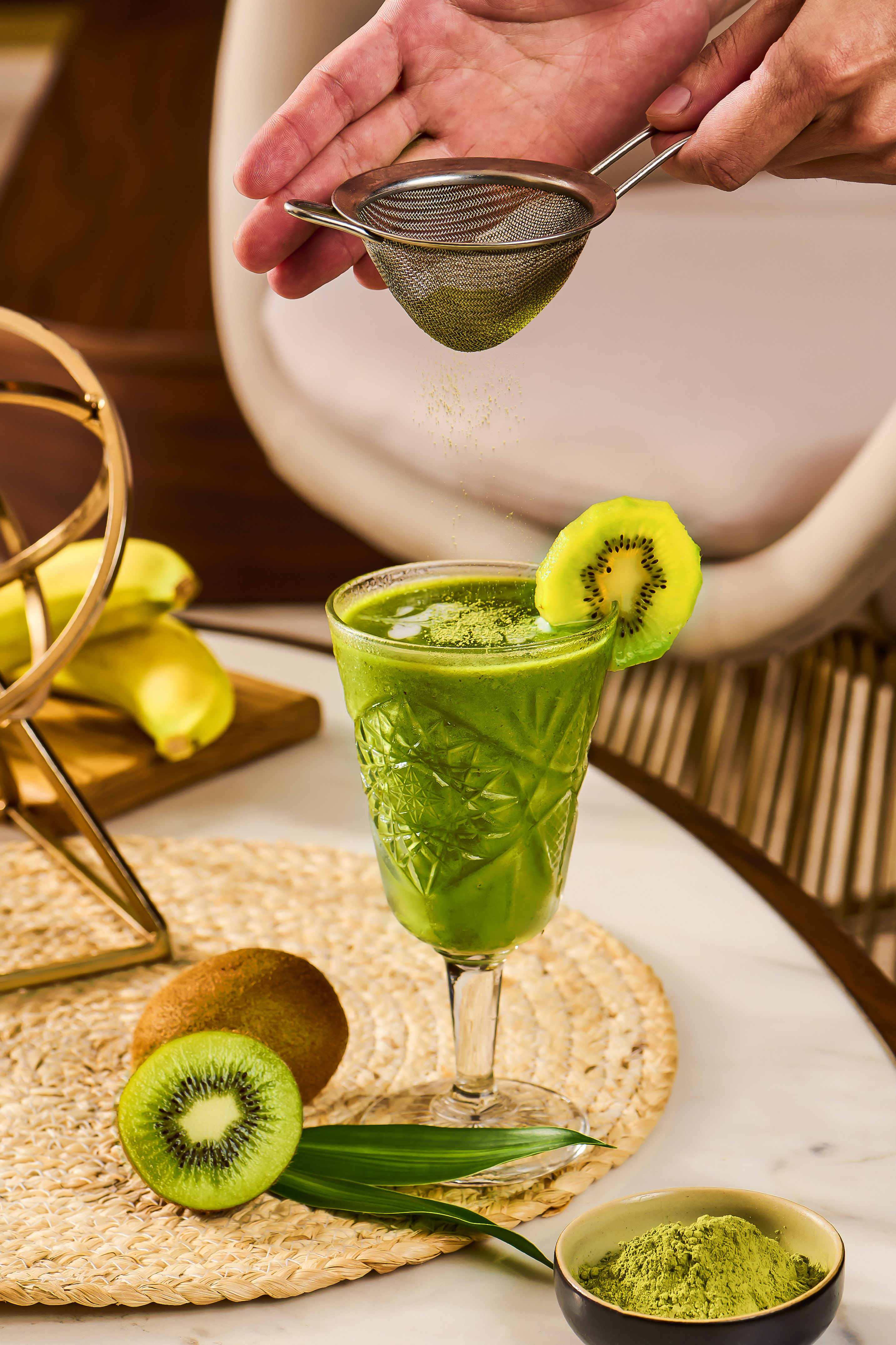 Hand sprinkling matcha powder over green kiwi smoothie with fresh kiwis and bananas.