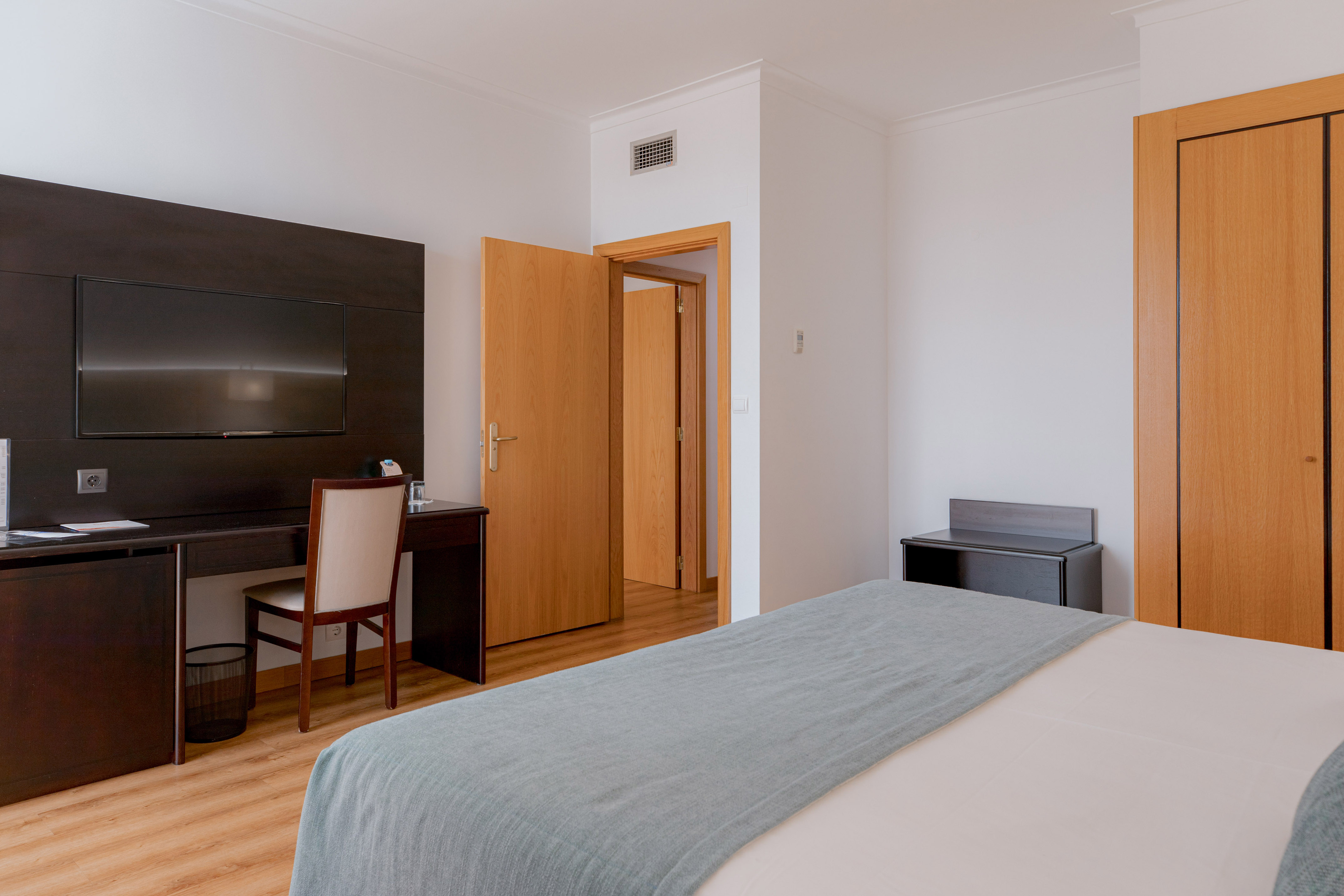 Modern hotel room with bed, desk, wall-mounted TV, and wooden flooring.