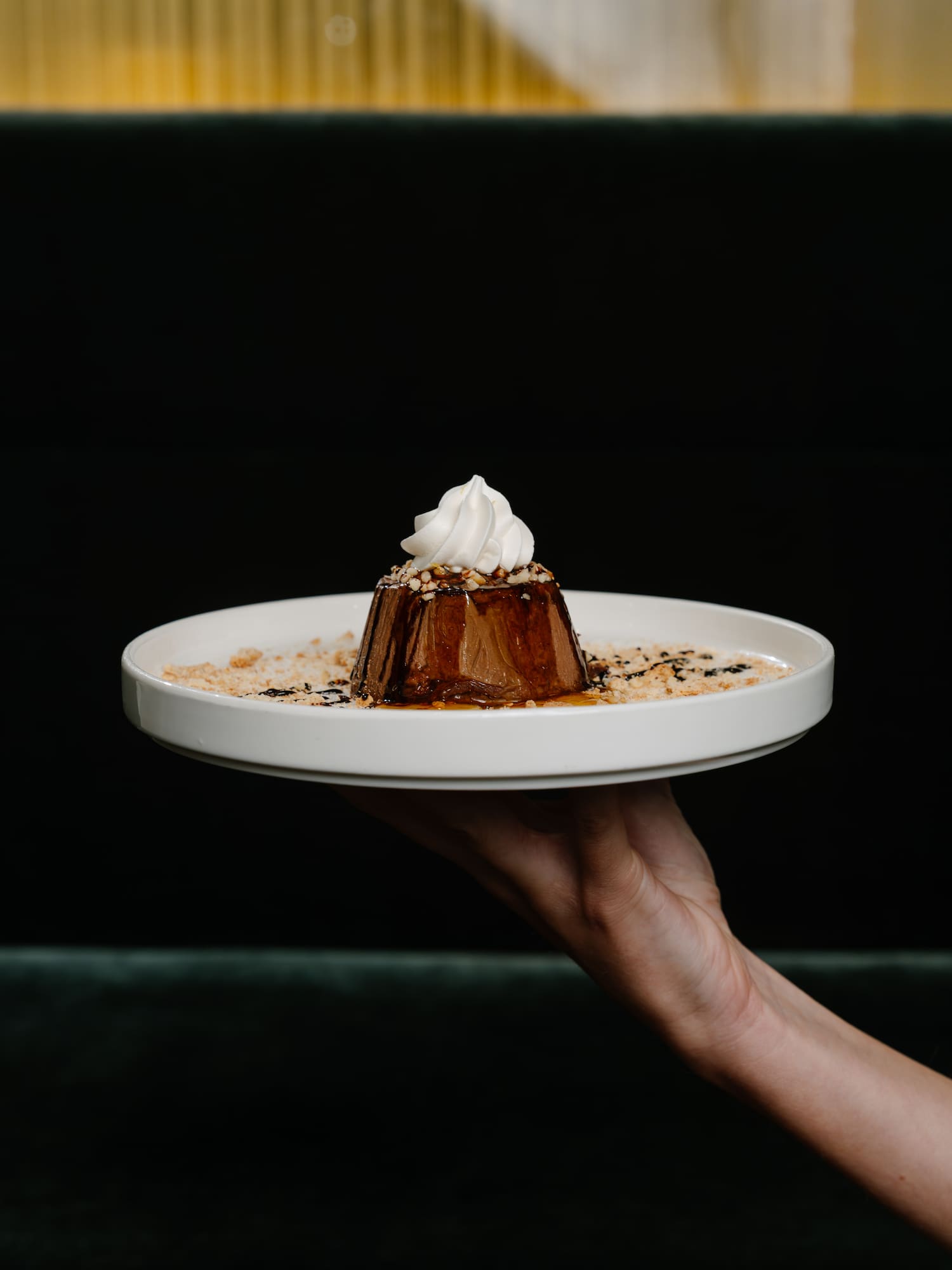 Hand serving chocolate dessert with whipped cream and nuts on white plate.