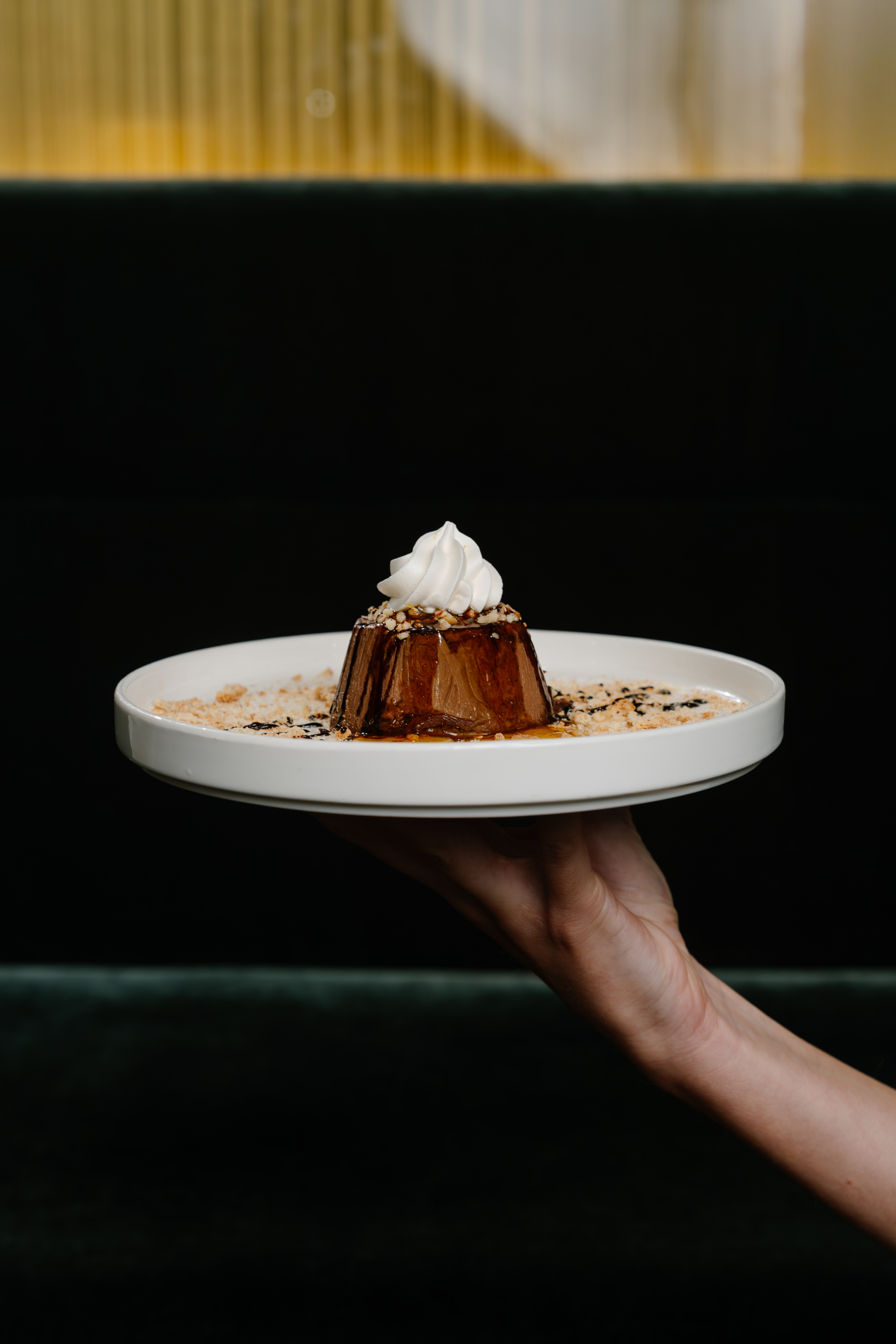 Hand serving chocolate dessert with whipped cream and nuts on white plate.
