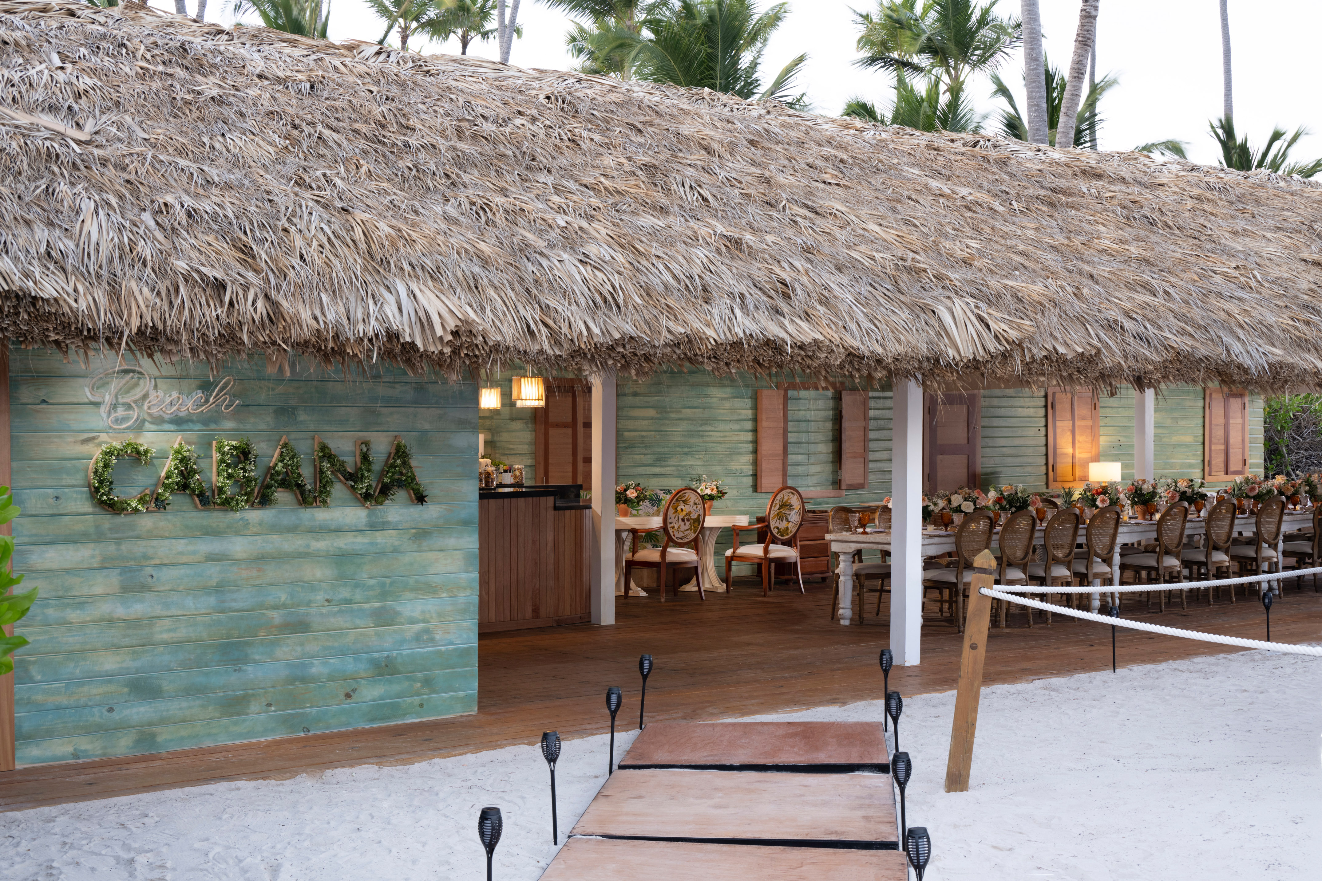 Thatched beach cabana, 'Beach Cabana' foliage sign, dining tables, sandy path.