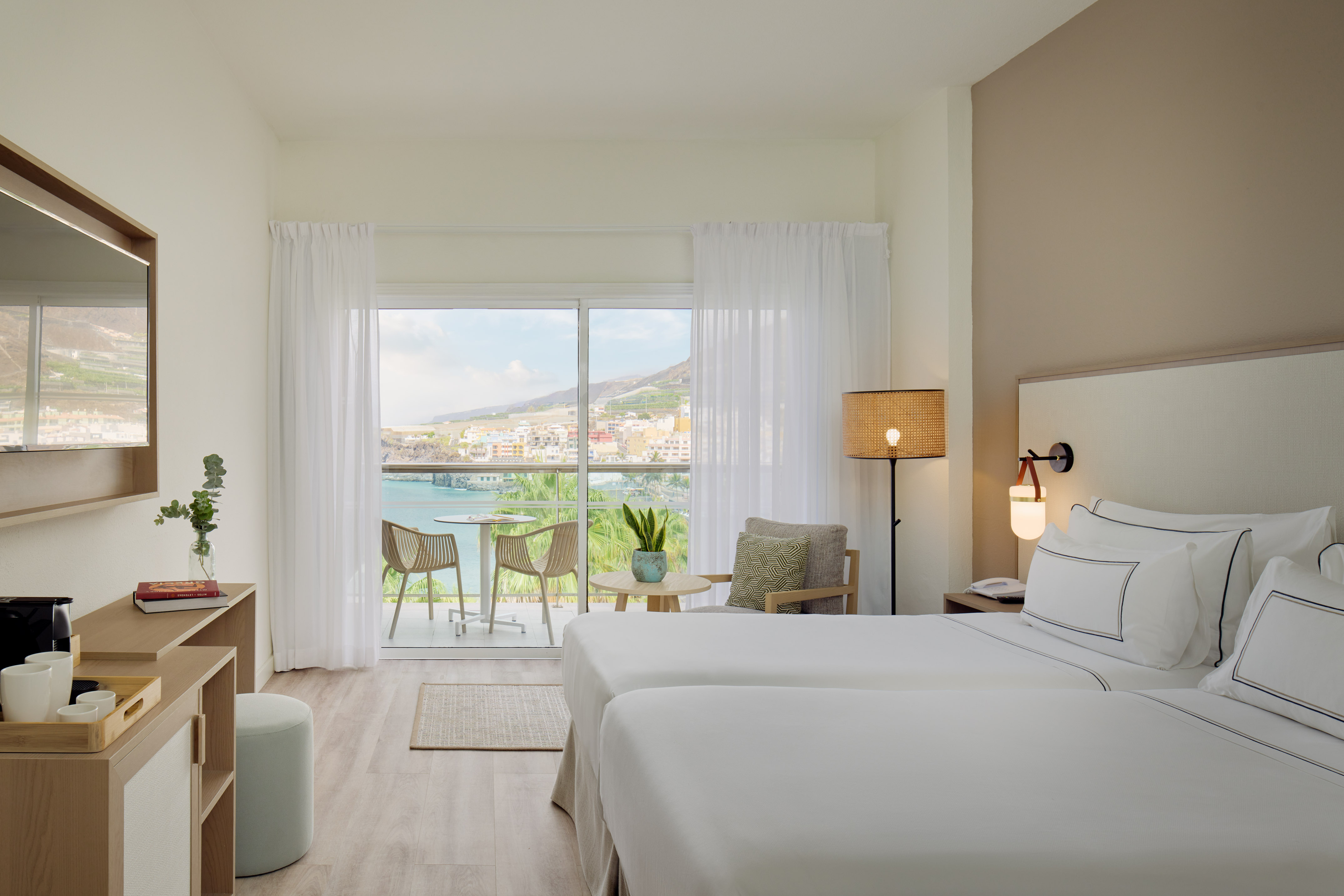 Hotel room with twin beds, balcony, and ocean view over coastal town.