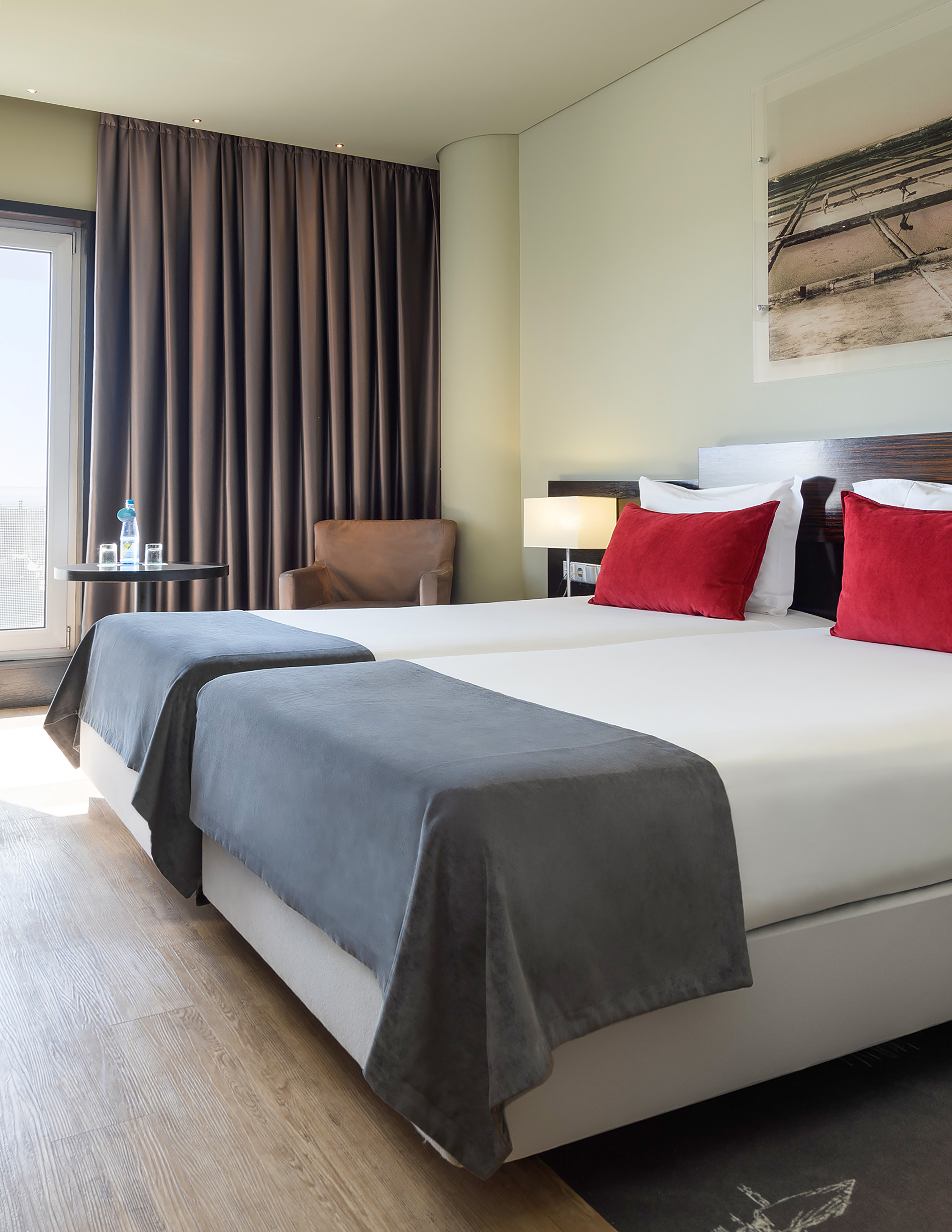 Modern hotel room with twin beds, red pillows, window, and wooden floor.