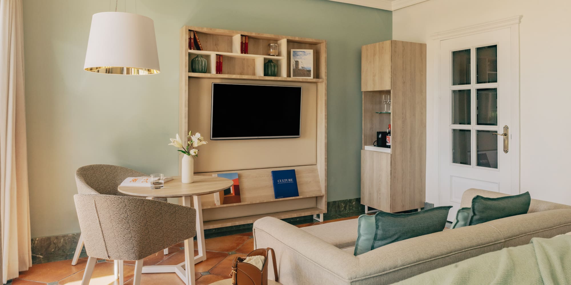 Hotel suite with green walls, terracotta floor, TV, comfortable sofa, and dining area.