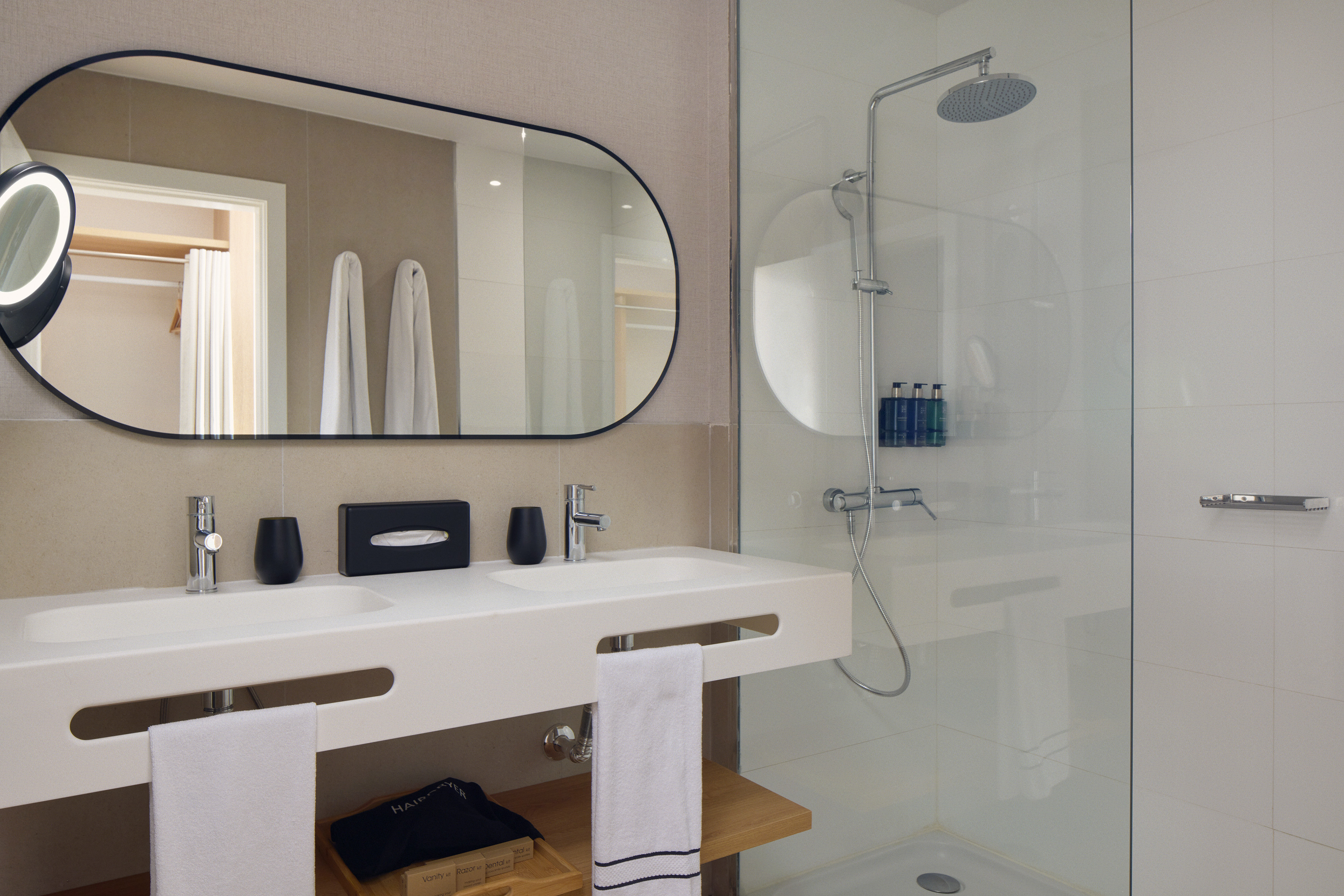 Modern hotel bathroom with double vanity, oval mirror, and glass shower.