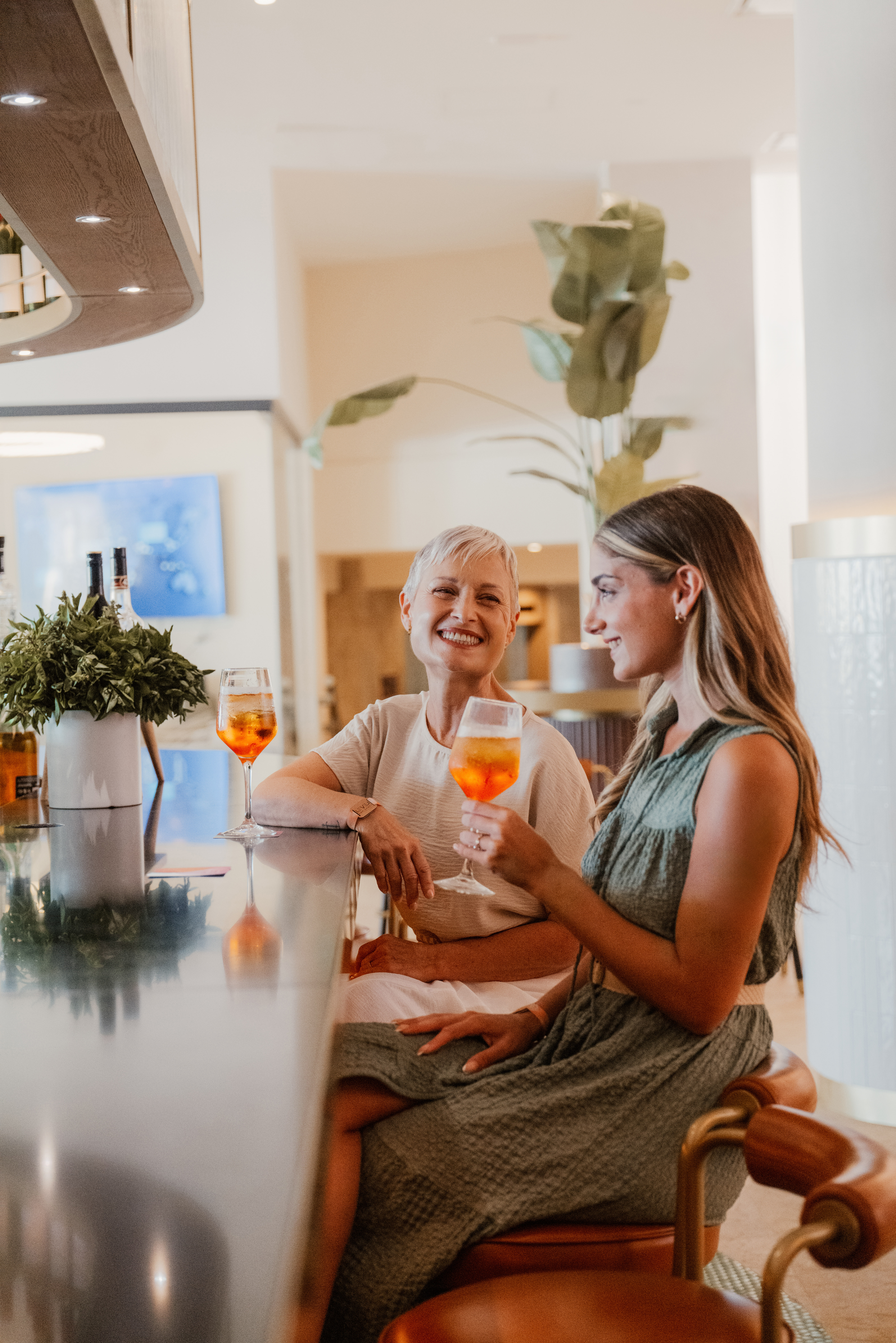 Two women smiling, enjoying Aperol Spritz cocktails at a modern bar.
