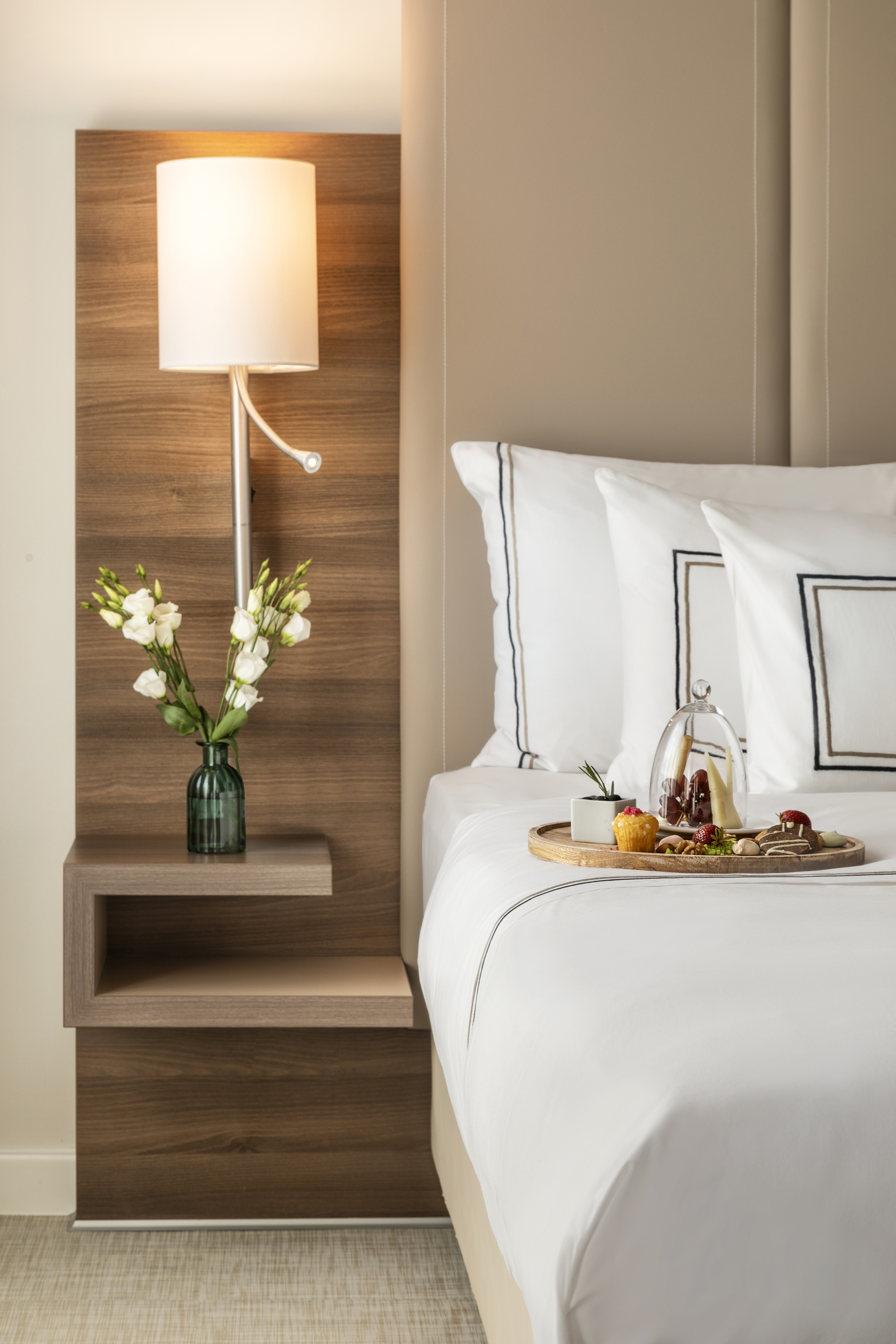 Luxury hotel bed with white linens, bedside lamp, flowers, and dessert tray.