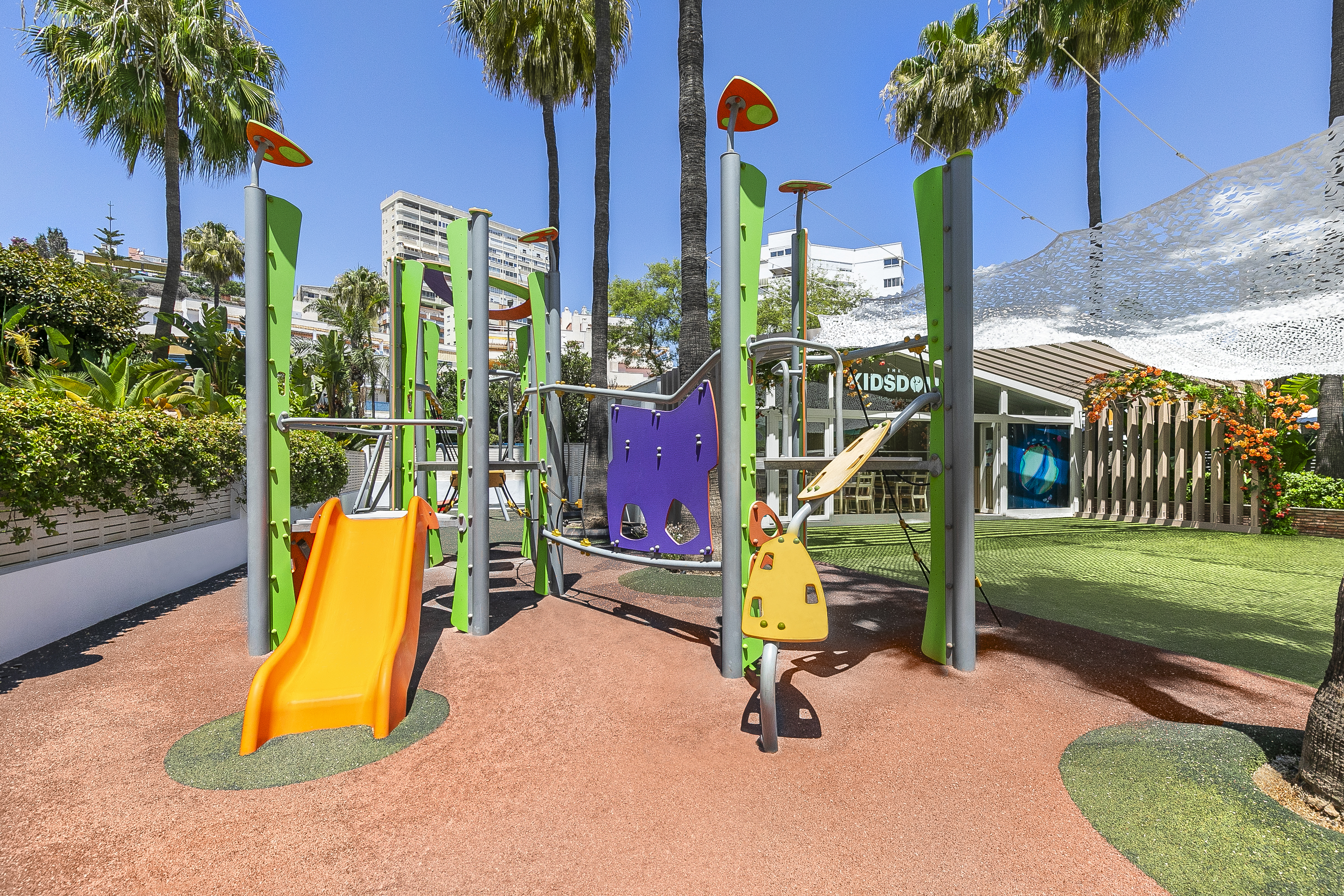 Outdoor resort playground with orange slide, climbing frames, and lush palm trees.