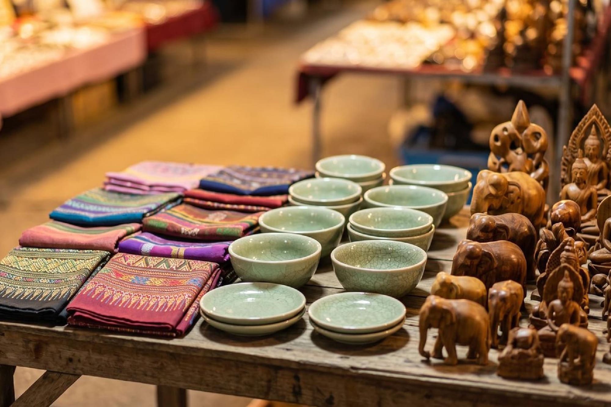 Market stall, patterned fabrics, green bowls, wooden elephant & Buddha carvings.