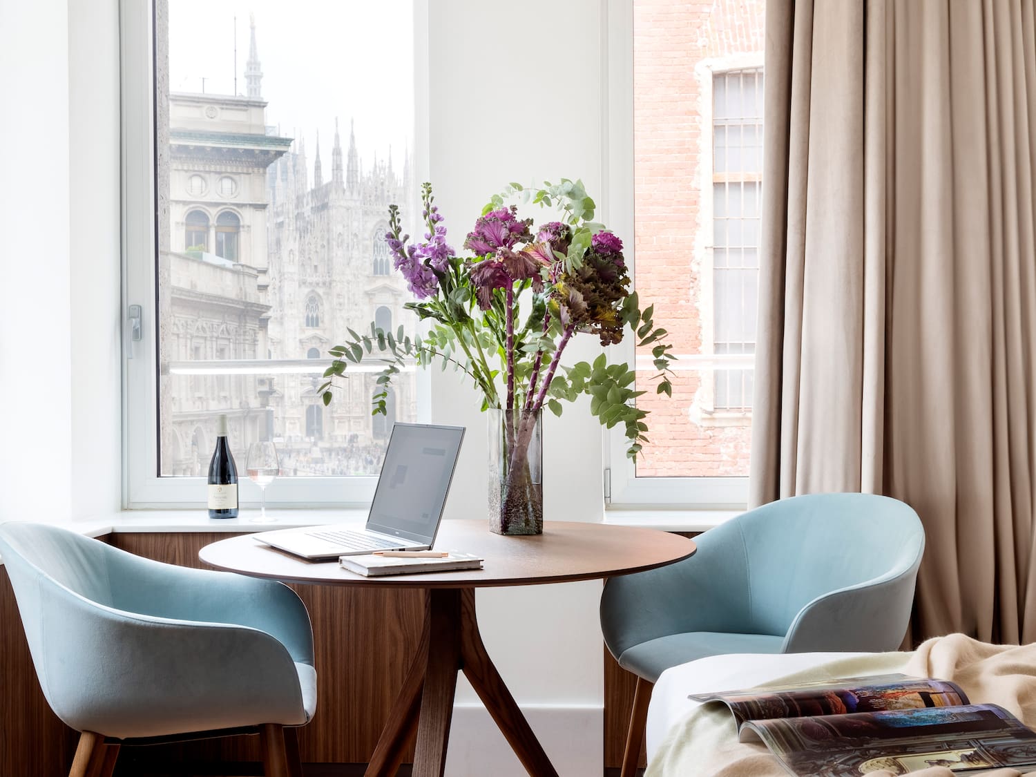 Milan Duomo view from room with round table, laptop, flowers, and blue chairs.