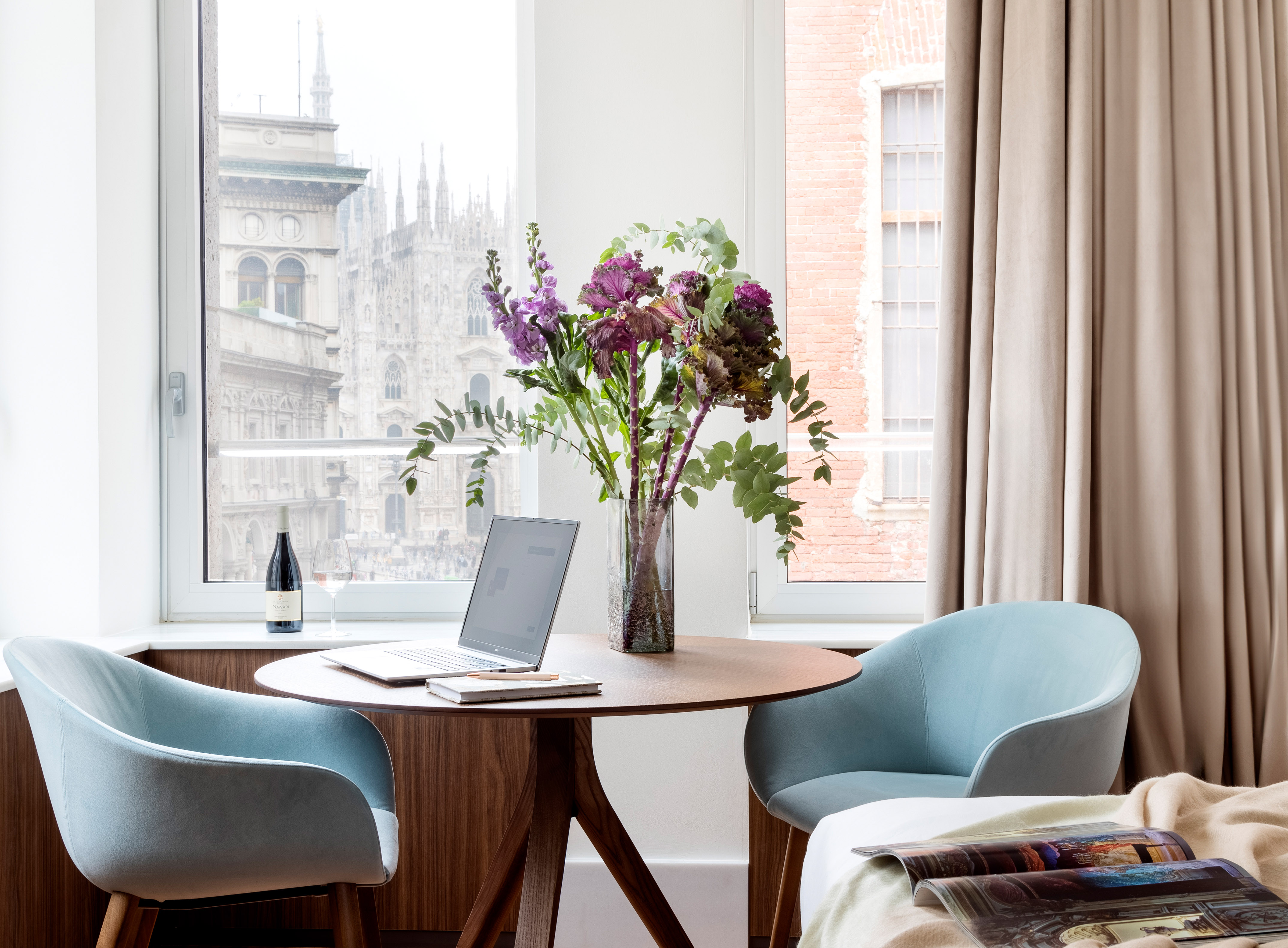 Milan Duomo view from room with round table, laptop, flowers, and blue chairs.