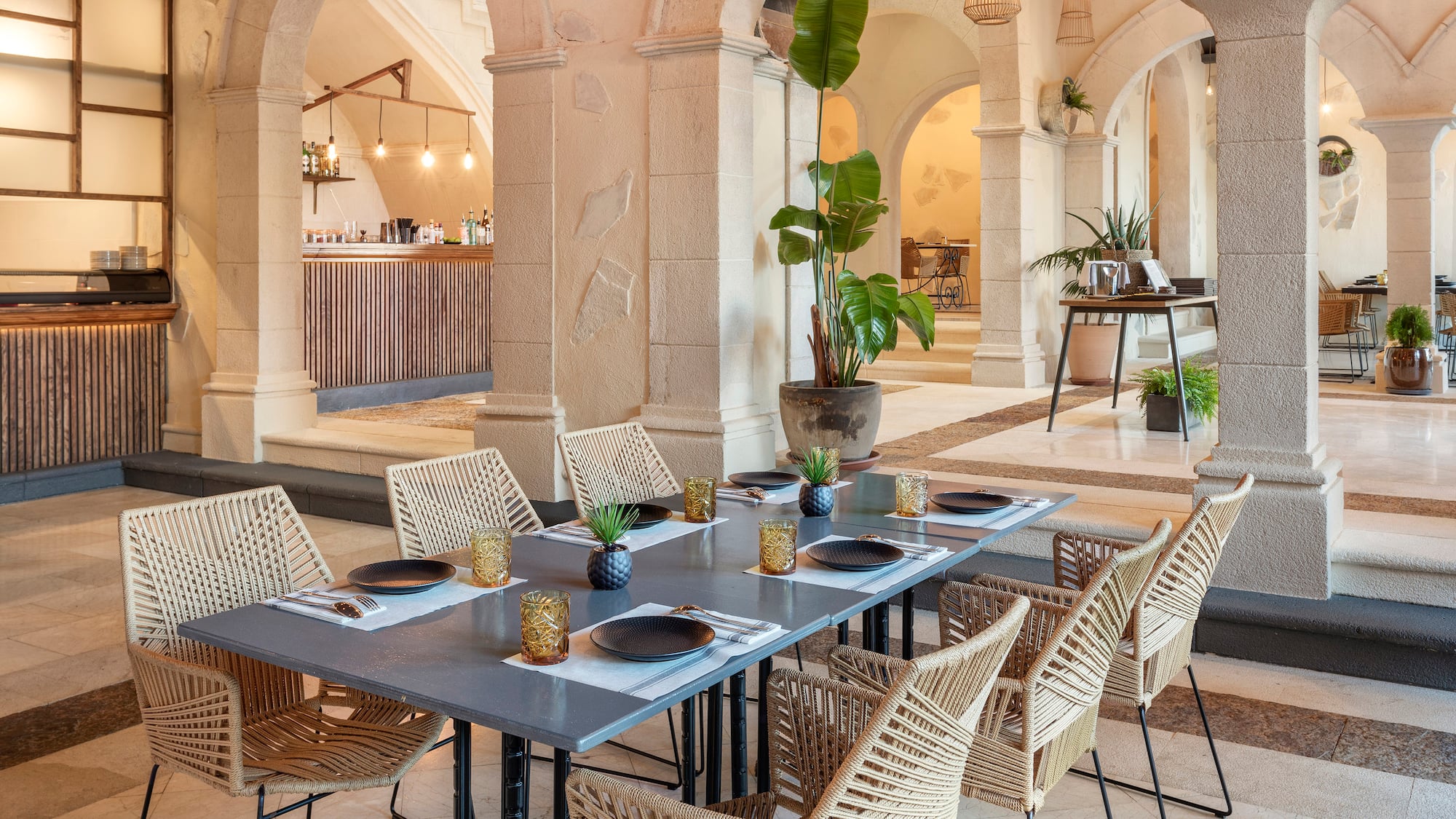a table with plates and wicker chairs in a room with columns and a bar