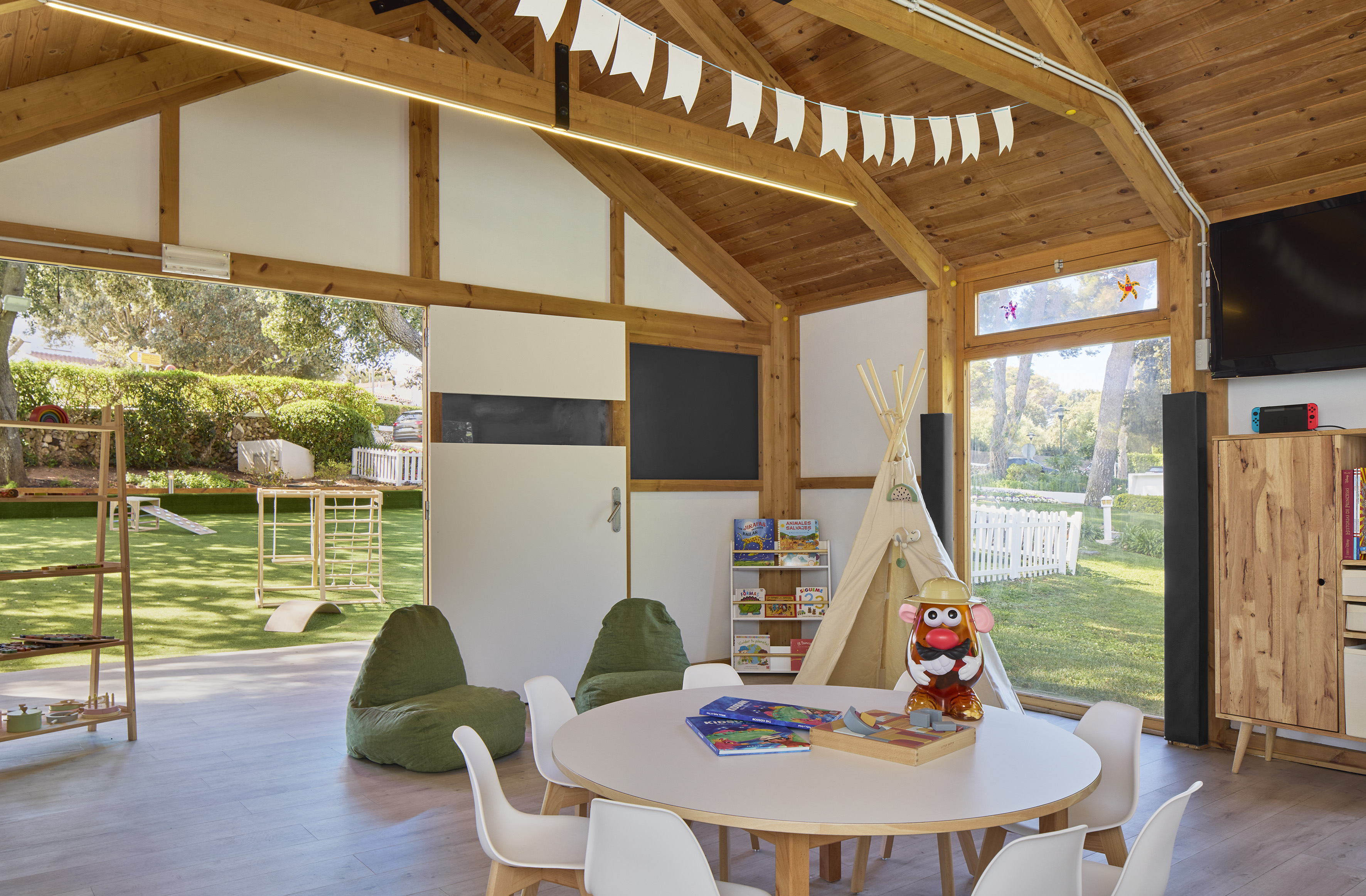 Vibrant kids' club with wooden beams, play table, teepee, toys, and outdoor garden.